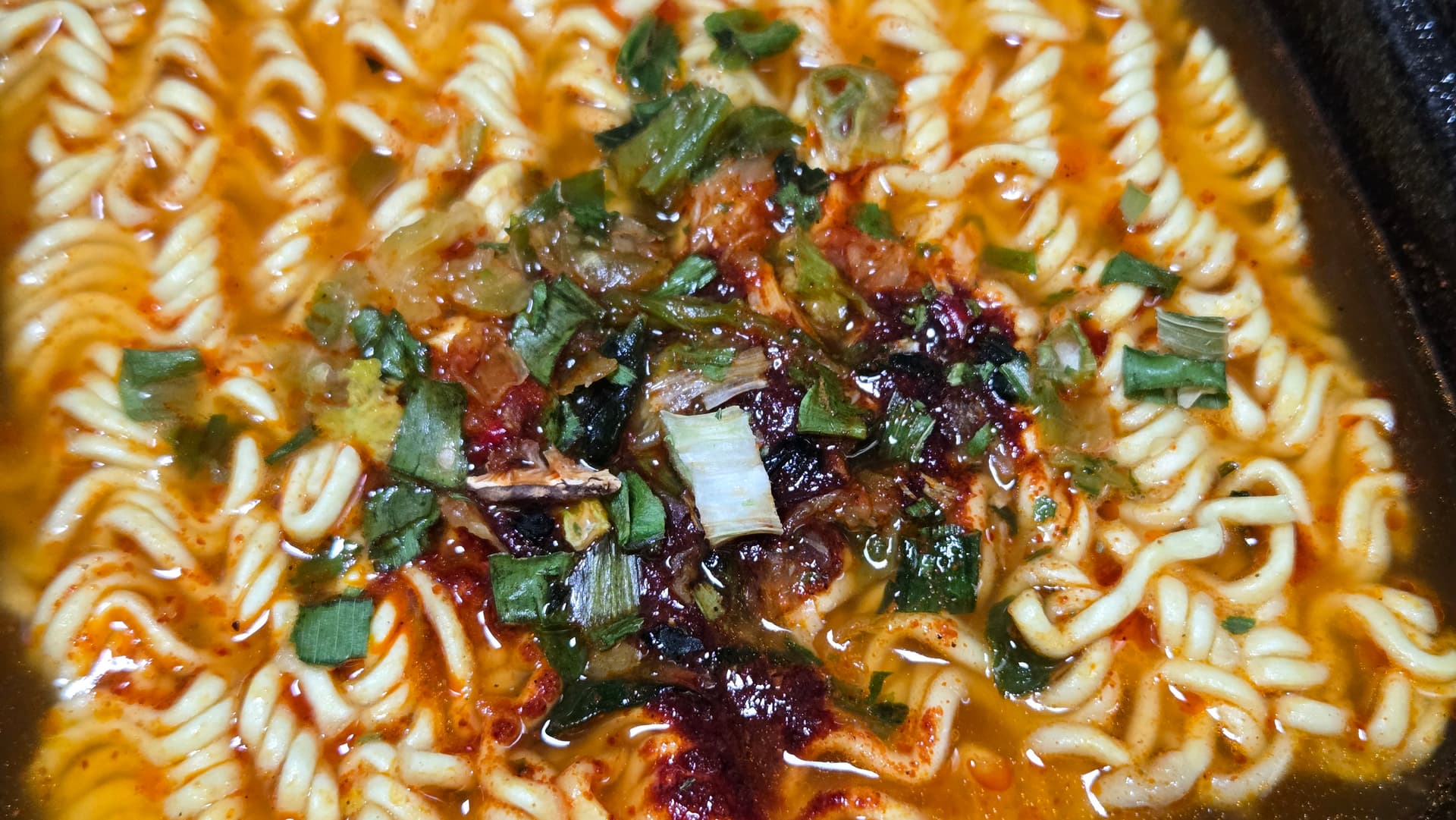 ຮູບໃກ້ມີ້ເຜັດເກົາຫຼີ ທຶມແຊລາມຽນ ປາລເກທັອກ - ເຫັນເມັດໝາກເຜັດແລະຜັກບົ່ວລອຍຢູ່ໃນນ້ຳແກງສີແດງເປັນມັນ | 하이제이에스비