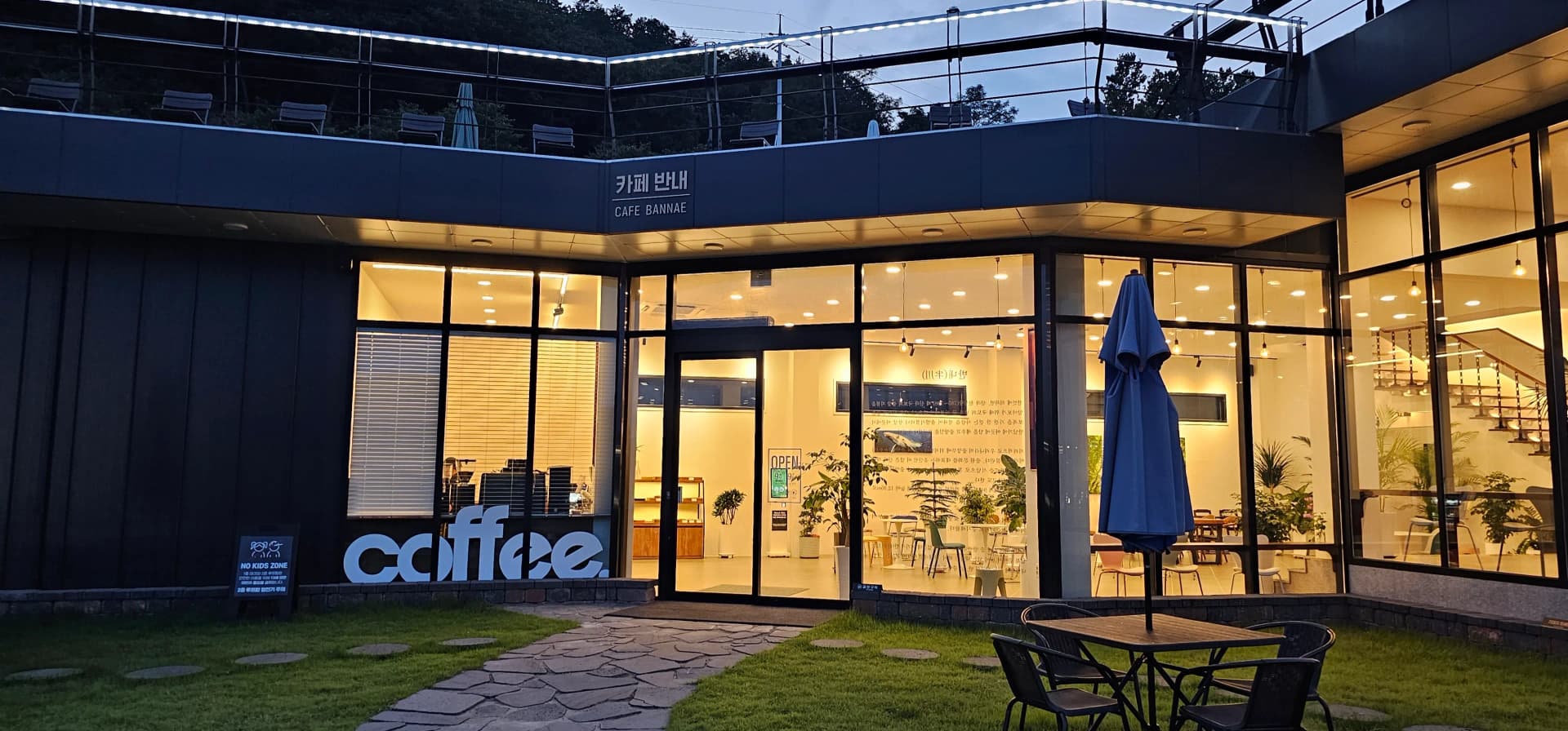 카페 반내 정면 — 대형 전면 유리와 잔디밭, 테라스 테이블이 보이는 저녁 풍경