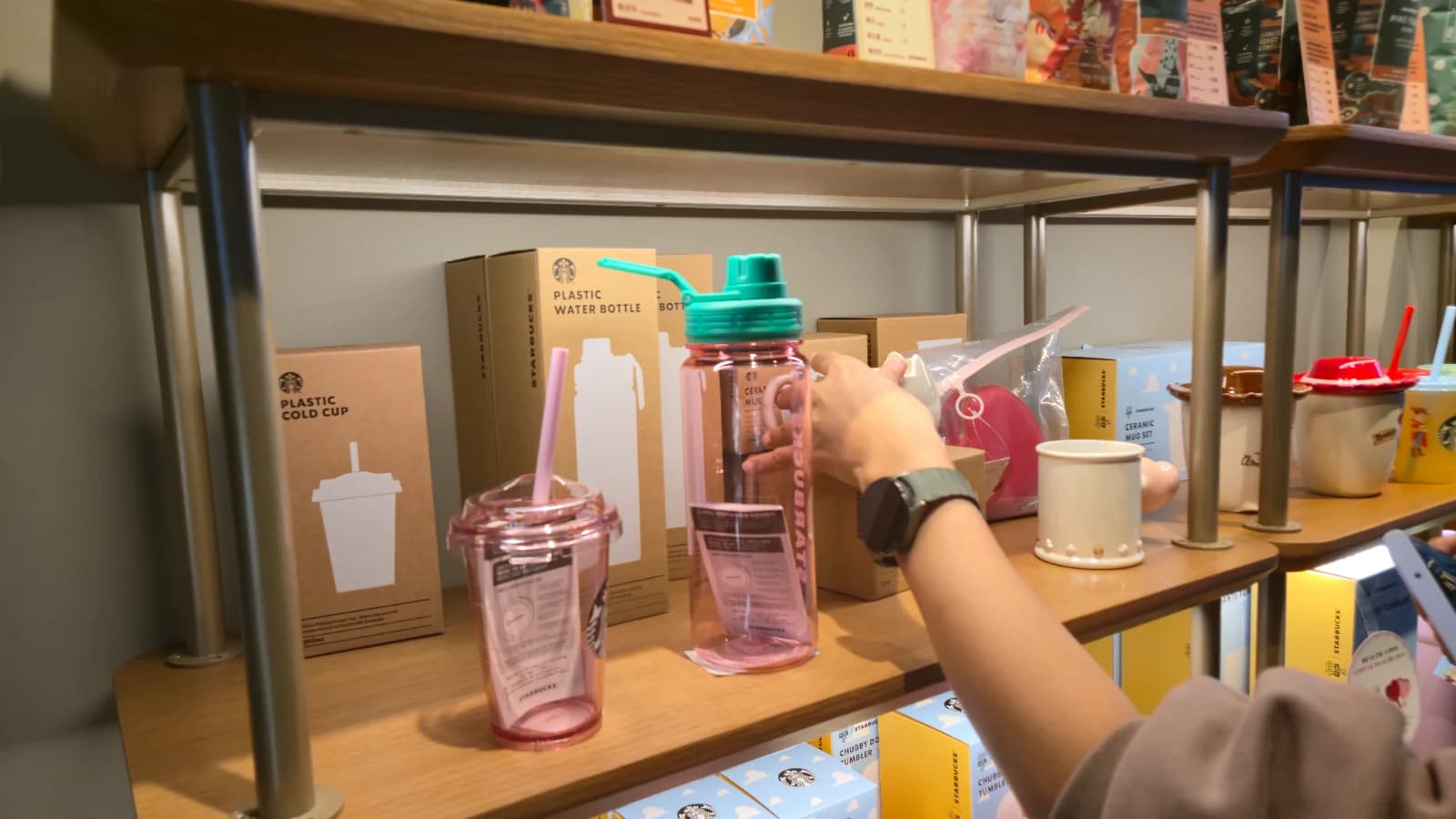Seção de produtos do Starbucks coreano com garrafas, cold cups, tumbler rosa e mão da minha esposa
