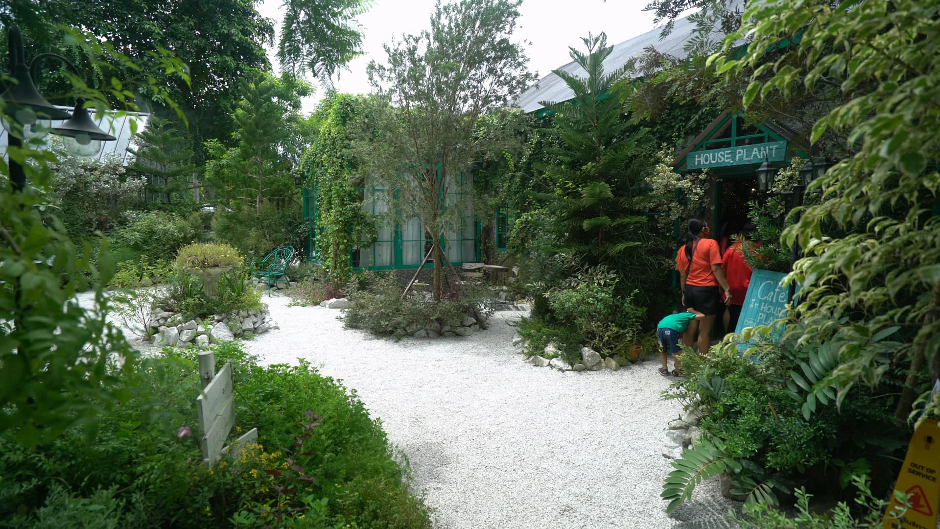 White gravel path lined with dense tropical shrubs and the HOUSE PLANT sign visible at the garden entrance