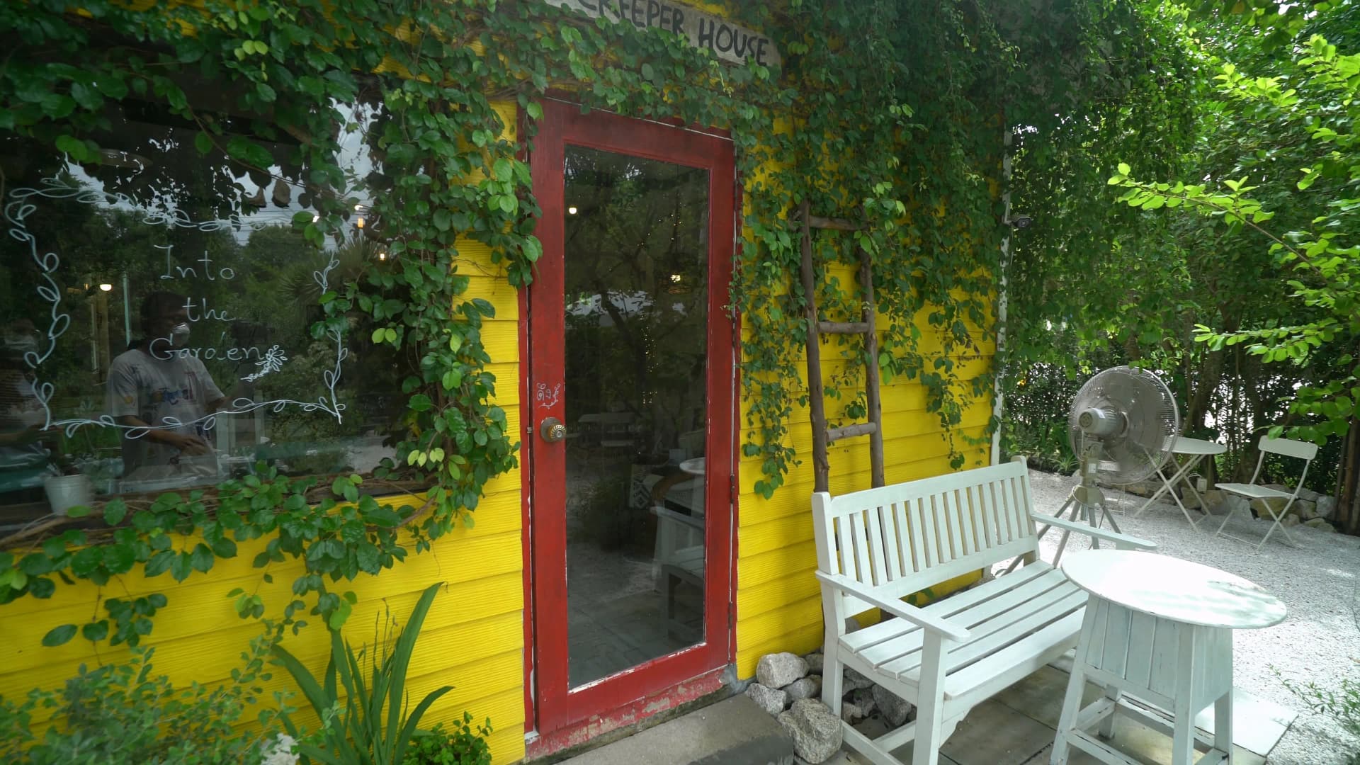 Building with yellow exterior wall and red door frame half-covered in vines with Into the Garden handwritten on the glass window