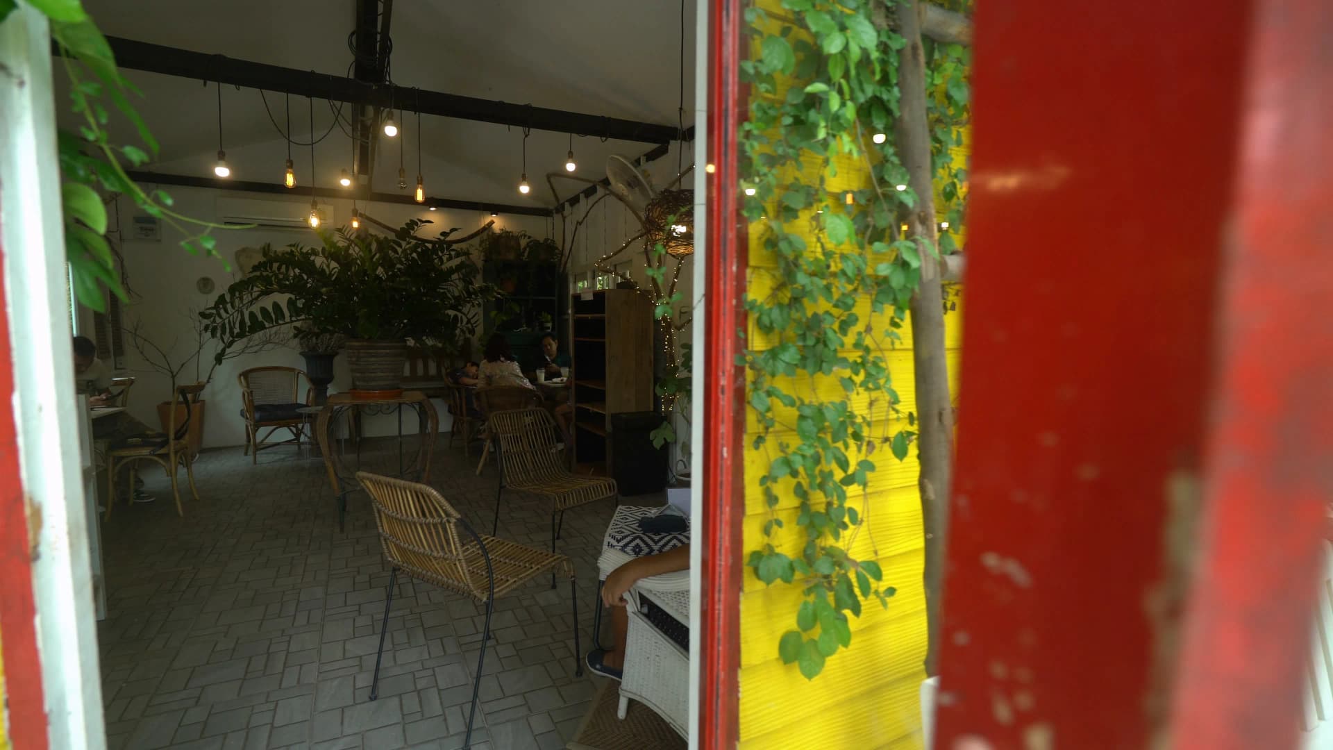Interior seen through the red door frame revealing rattan chairs, Edison bulb string lights, and a large potted plant