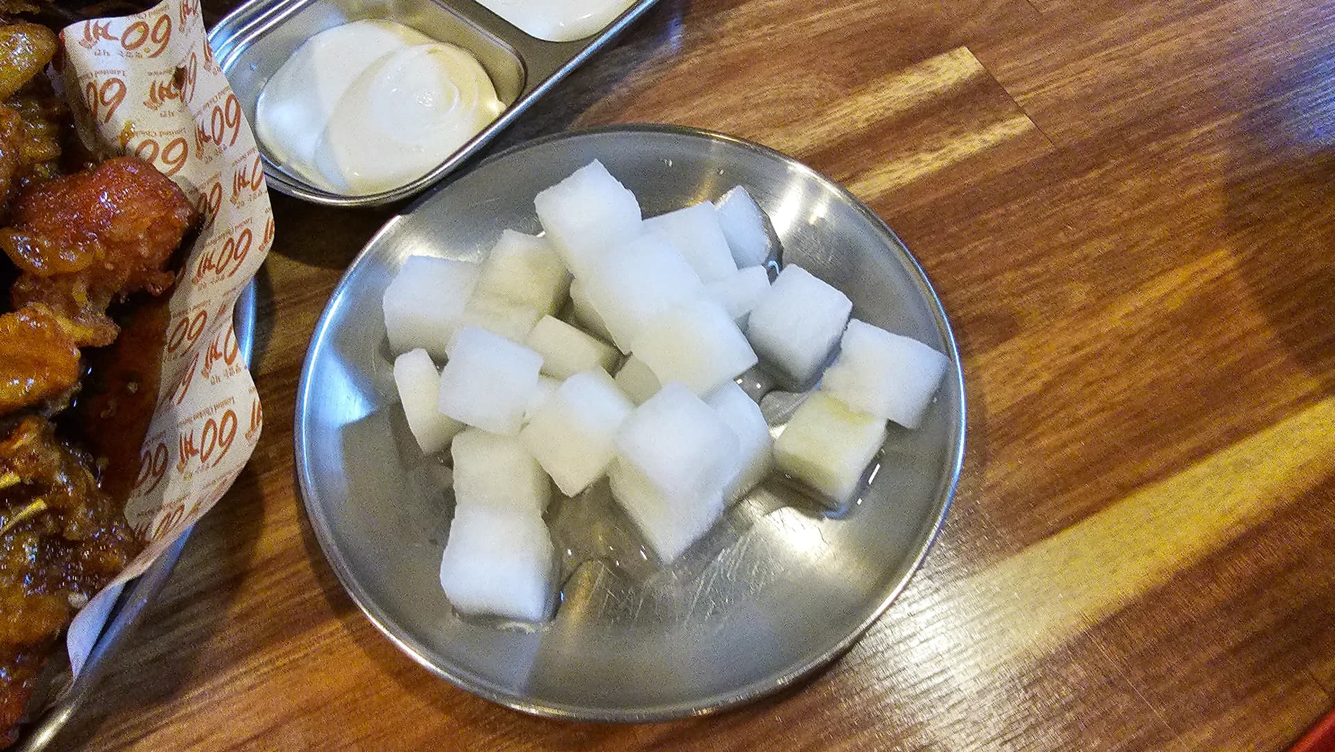 وعاء ستانلس ستيل فيه مكعبات فجل أبيض مخلل شفاف بجانب طبق دجاج مقلي كوري