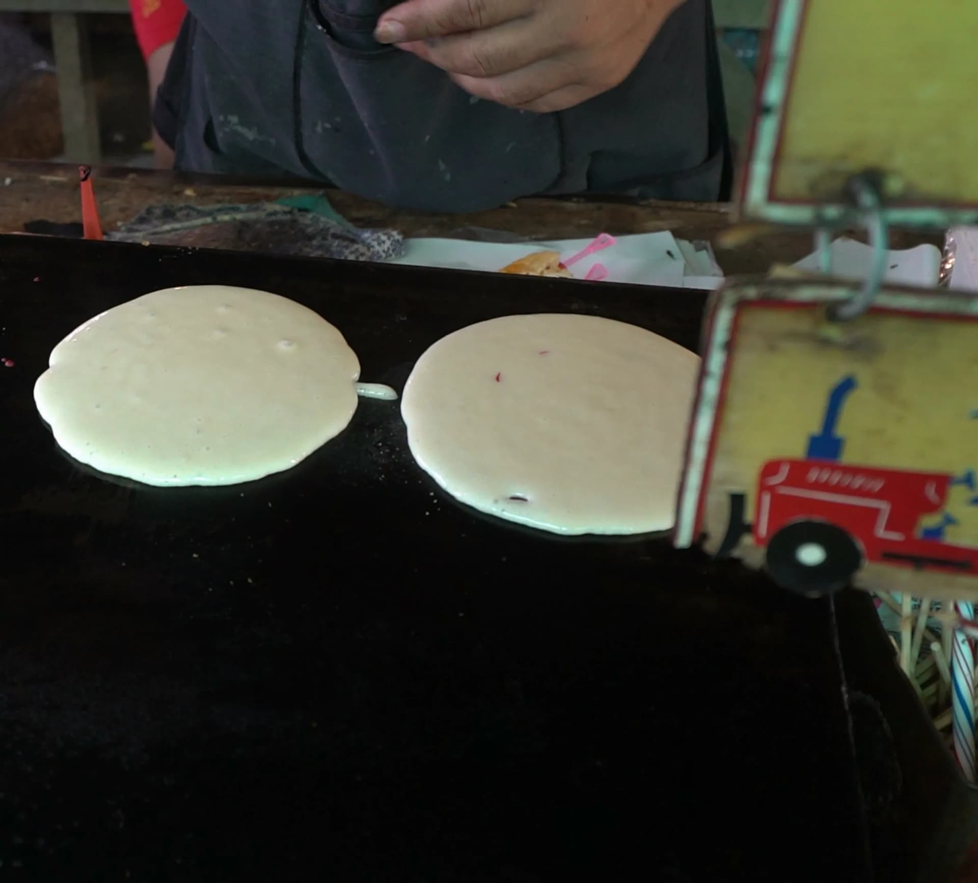 태국 시장 팬케이크 만드는 모습 – 검은 철판 위에 하얀 반죽 원 두 개가 익어가는 장면