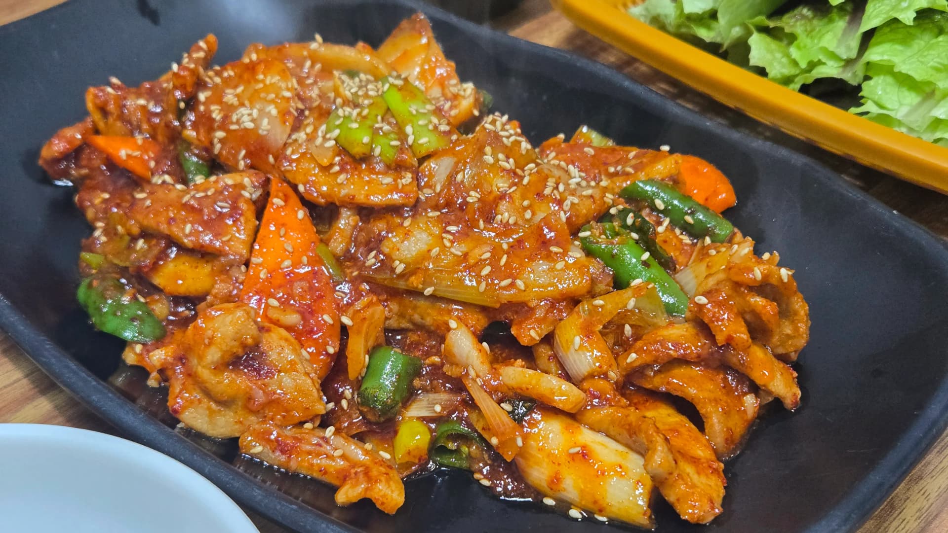 제육볶음 전체 모습 - 검은 철판 위에 고추장 양념으로 볶은 돼지고기 양파 대파 청양고추 당근과 쌈채소 바구니 | 하이제이에스비