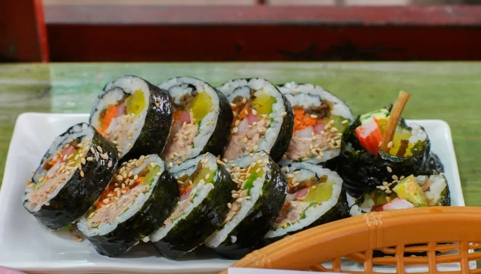 White plate full of tuna kimbap cross-sections sprinkled with sesame seeds | 하이제이에스비