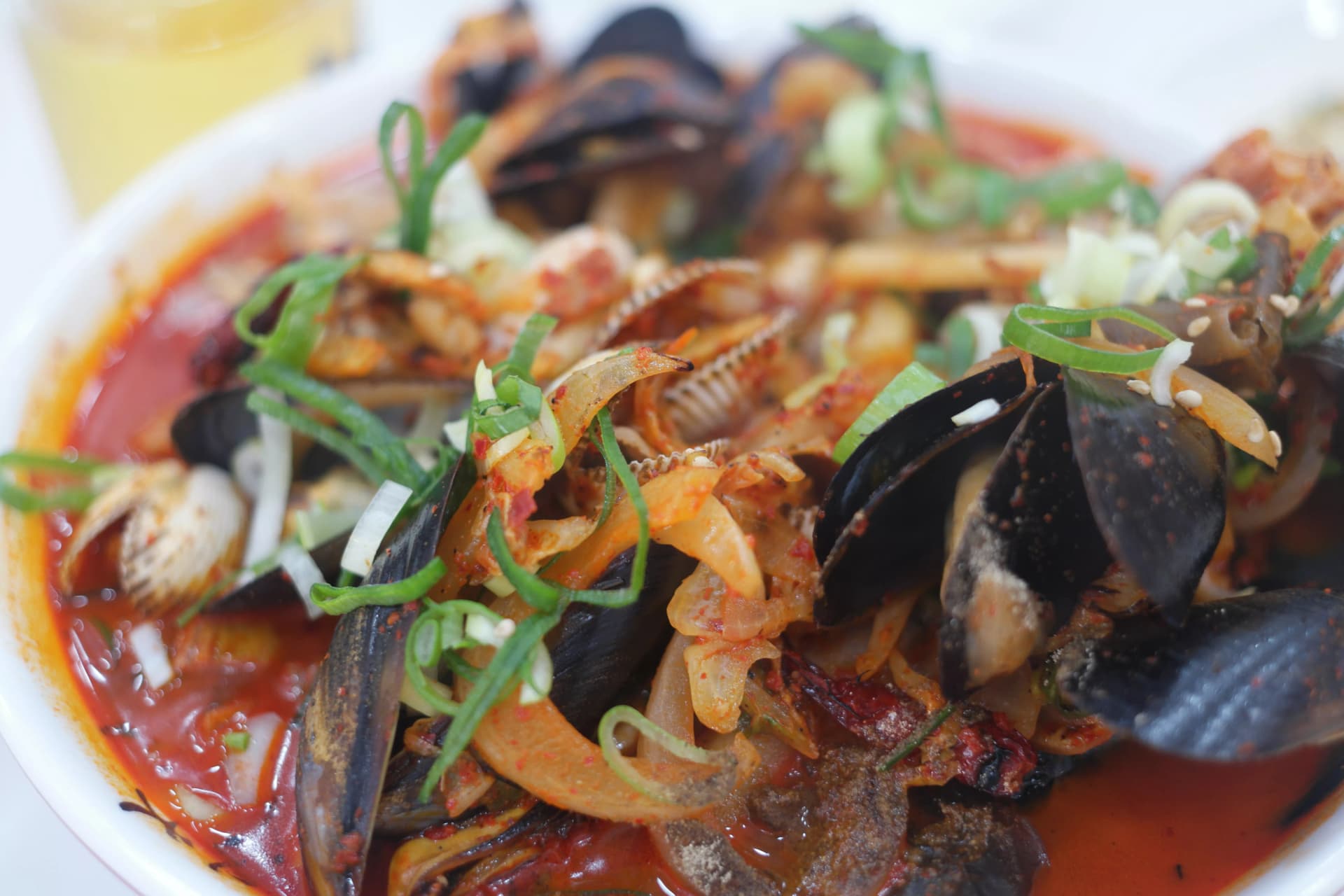 짬뽕 국물 클로즈업, 홍합 바지락 오징어가 진한 주홍빛 국물에 담겨 있고 고춧가루가 국물 위에 떠 있는 모습 | 하이제이에스비