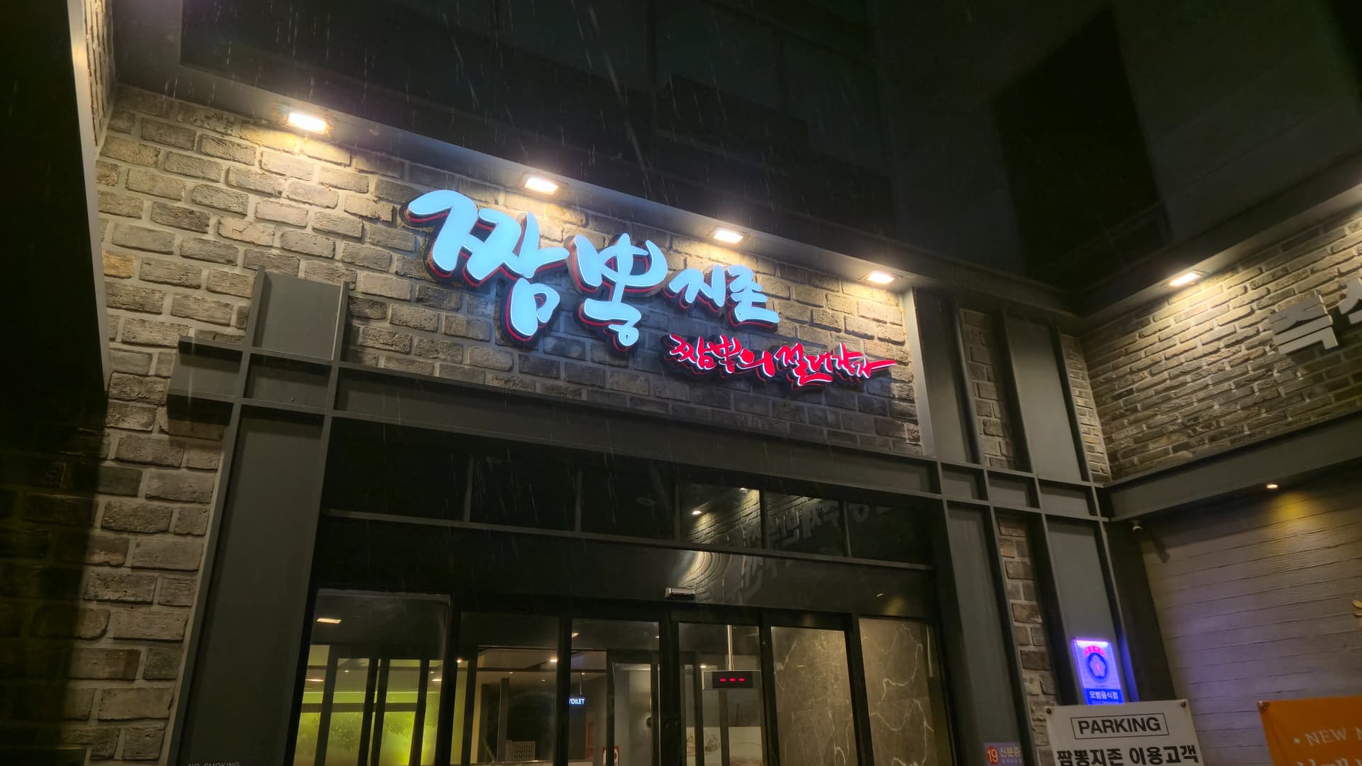 비 오는 밤 짬뽕지존 대전둔산점 외관, 파란색과 빨간색 간판이 켜져 있고 오른쪽에 주차 안내판이 보인다