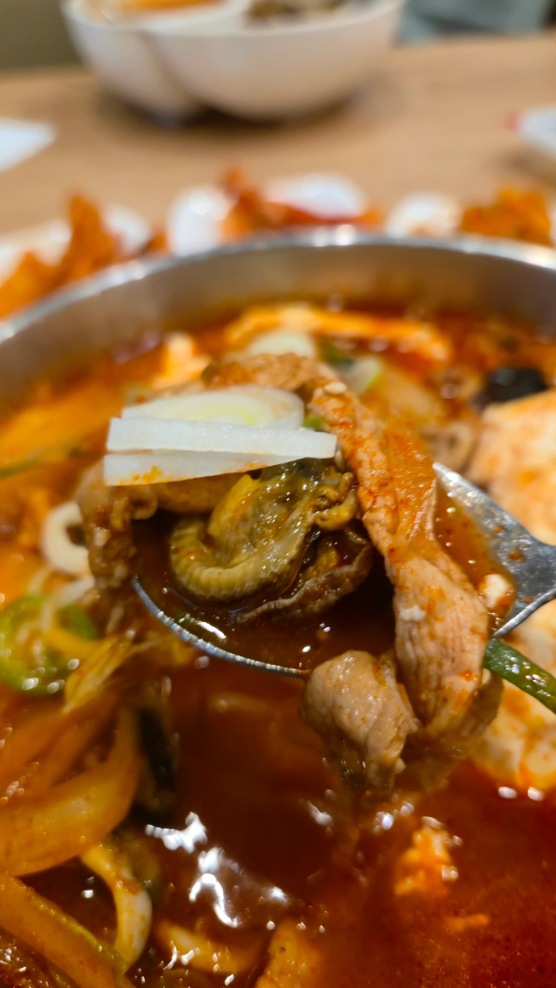 숟가락 위에 바지락 조개와 돼지고기가 올라와 있고 뒤로 빨간 짬뽕 국물이 보인다