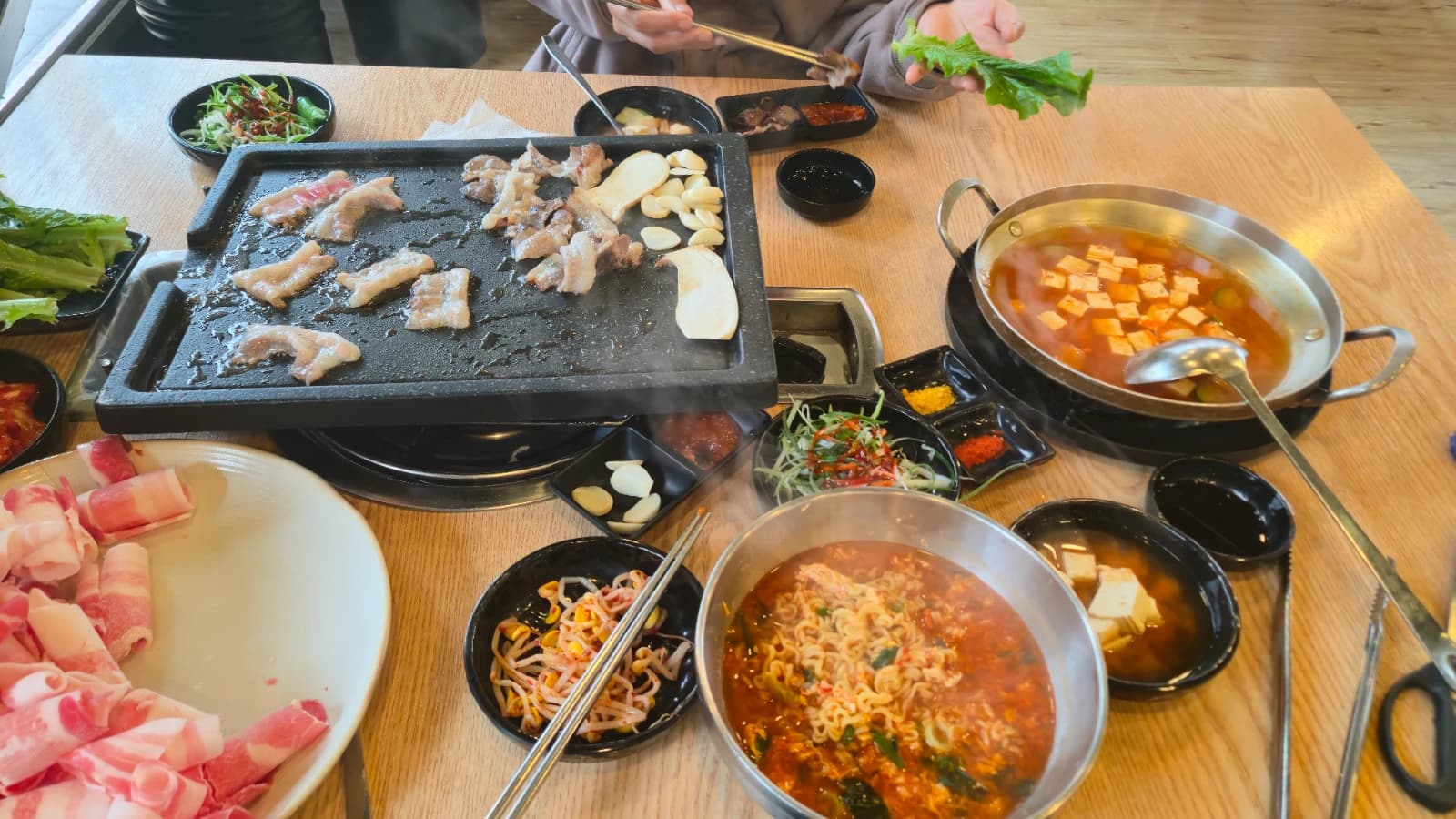 Mesa completa con daepae samgyeopsal asándose, sopa de doenjang, ramen y banchan