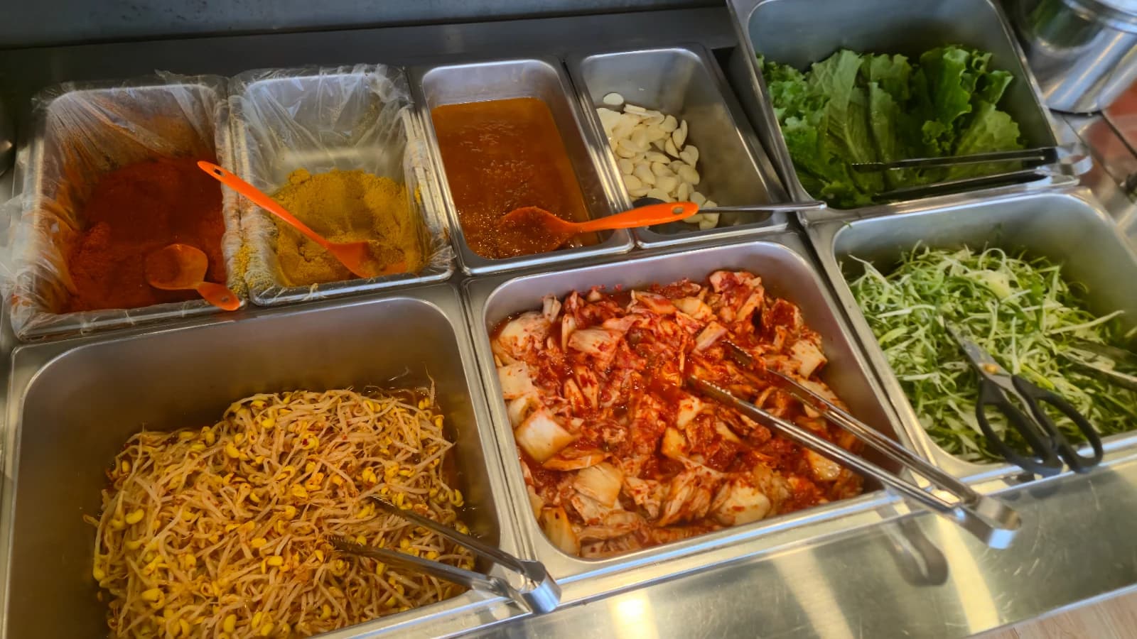 Vista completa del mesón self-service con kimchi, brotes de soja, lechuga, cebollín, ajo y salsas