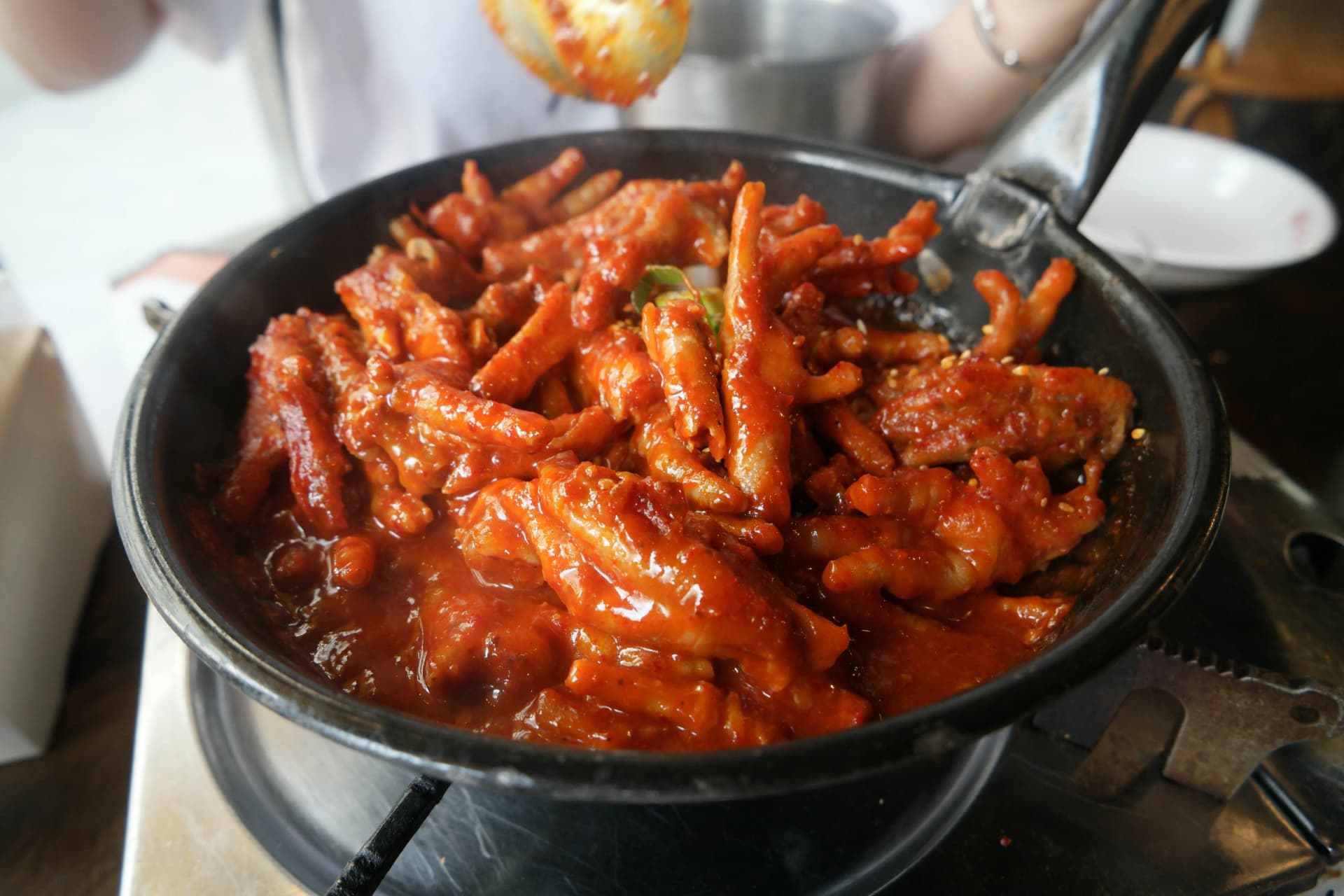 Patas de pollo picantes cocinándose sobre fuego de gas mientras las remueven con cucharón, con la salsa roja hirviendo a borbotones