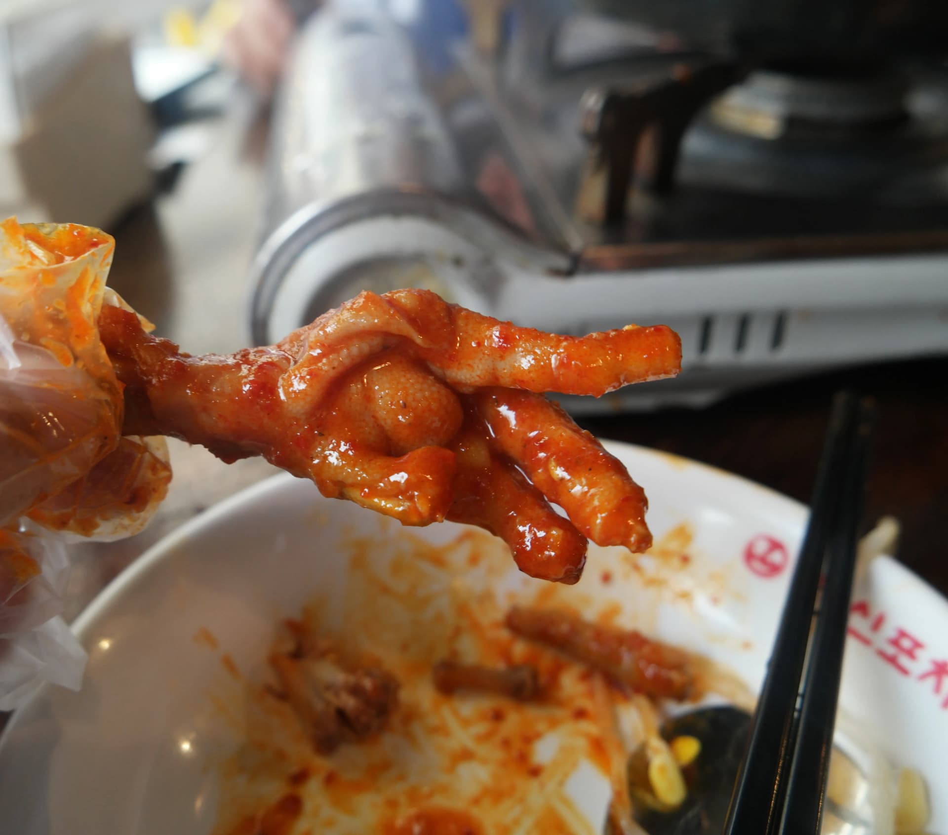 Una mano con guante plástico sosteniendo una pata de pollo picante, con la forma de los dedos claramente visible