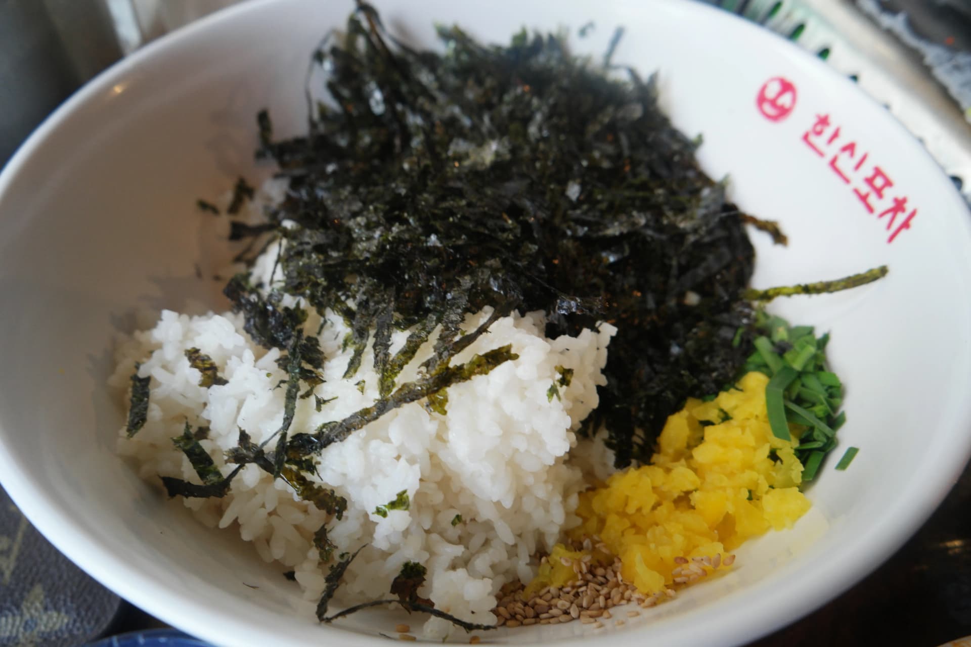 Ingredientes de jumokbap en bowl blanco con arroz, hojuelas de alga, rabanito amarillo encurtido, ajonjolí y cebolla china, con el logo de Hanshin Pocha