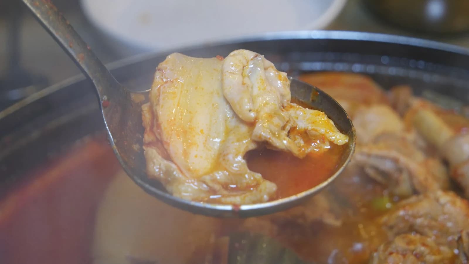 Ladle lifting a whole chicken drumstick from the stew with spicy broth dripping off and meat separating from the bone