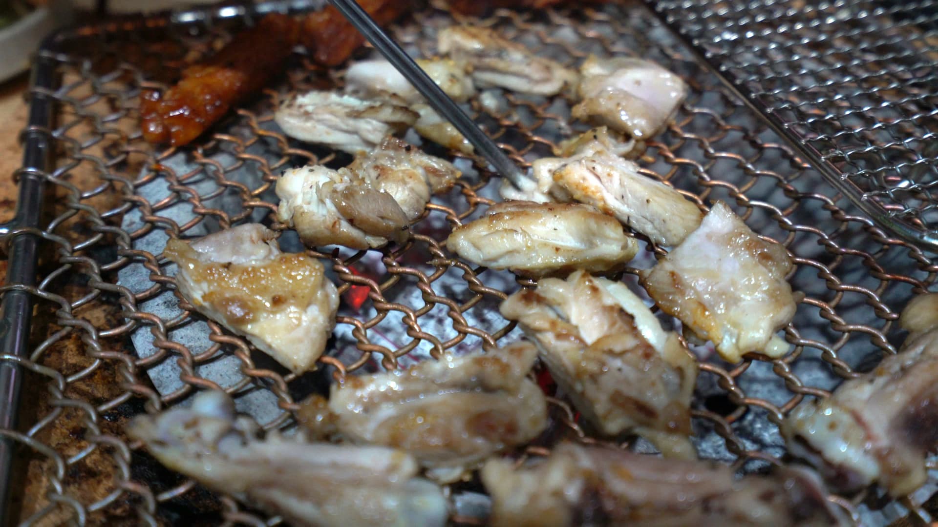 Salt-seasoned chicken turning golden brown on a charcoal mesh grate being flipped with tongs