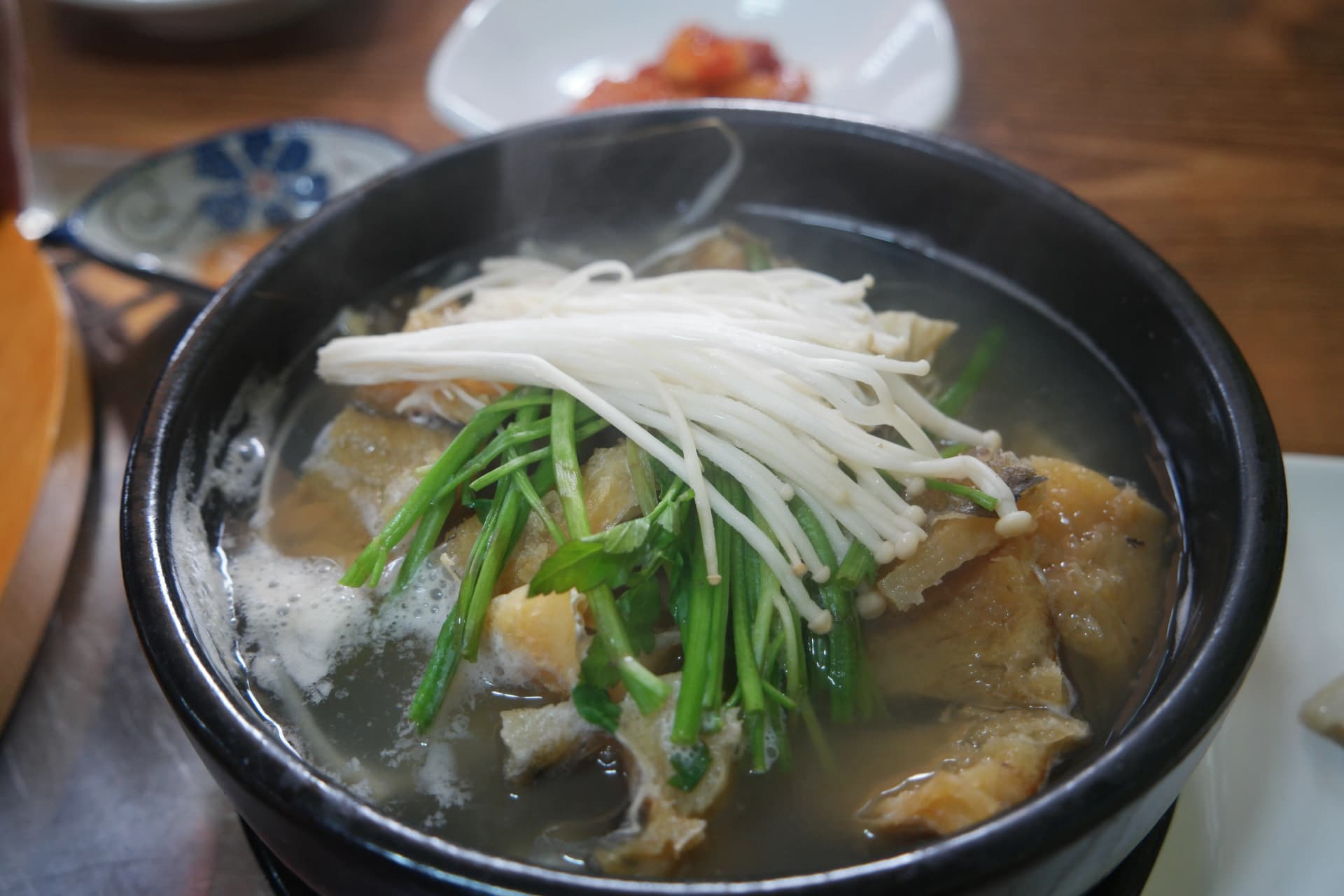 شوربة هوانغتيه صافية في وعاء فخاري أسود مع فطر إينوكي وكراث أخضر