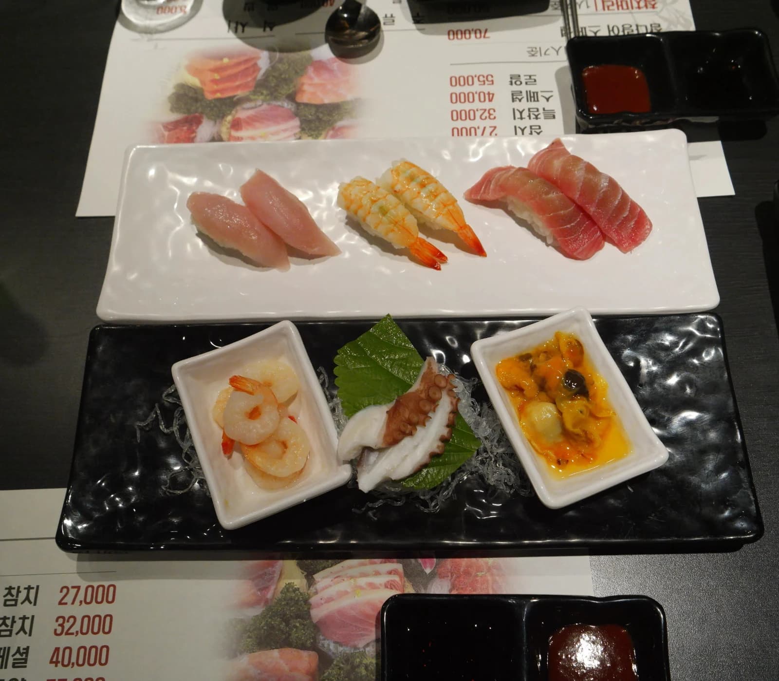Full table spread at a Korean sashimi restaurant with sushi on white plates and seafood side dishes on dark plates
