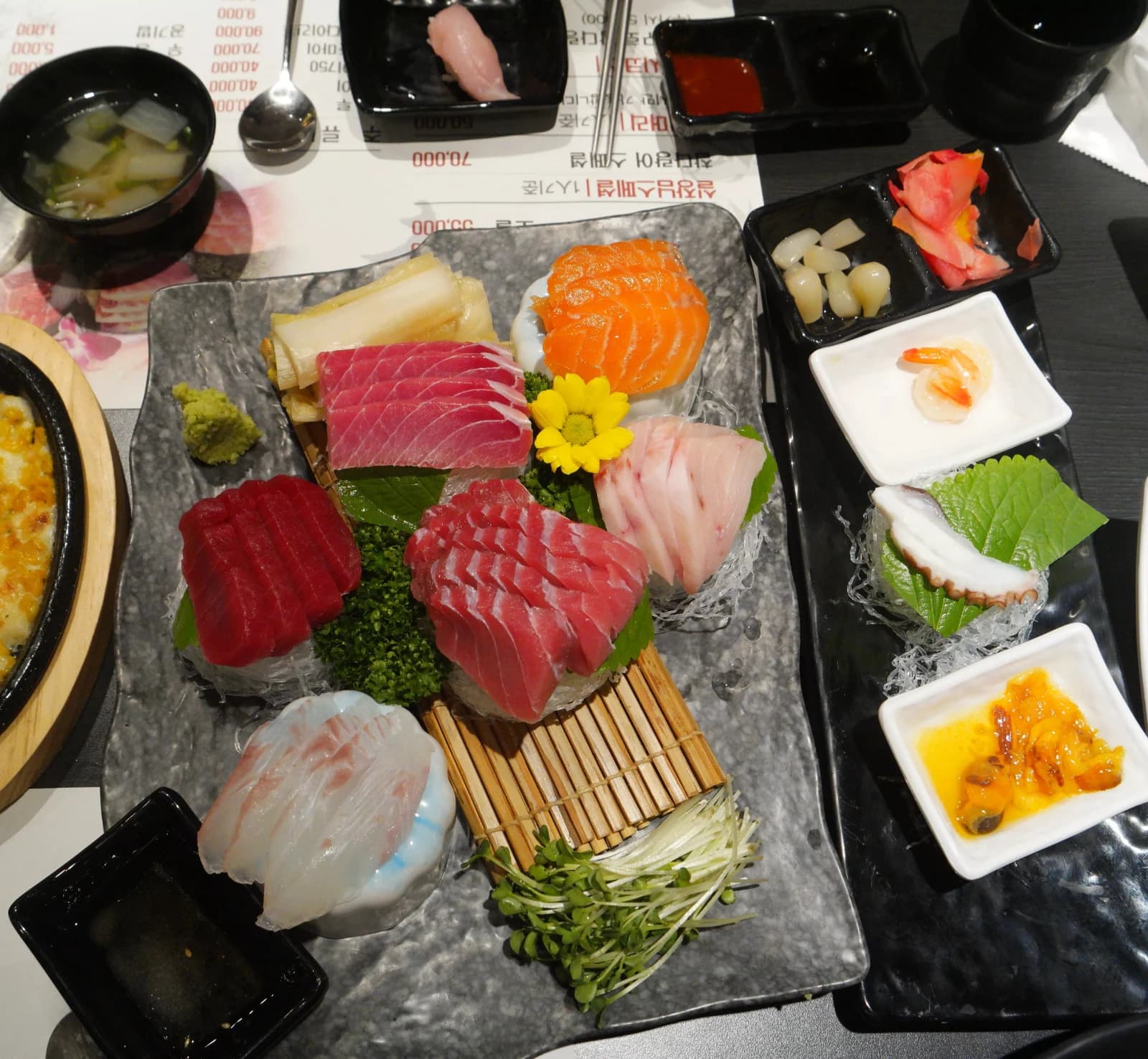 Full sashimi platter with multiple tuna cuts, flounder sashimi on bamboo, wasabi, and a chrysanthemum garnish surrounded by side dishes