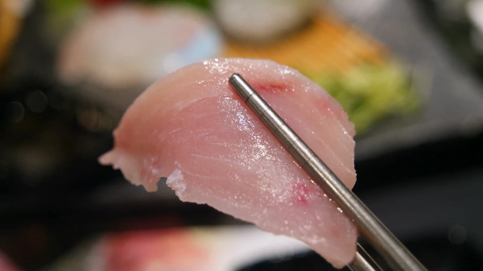 Chopsticks holding a thin translucent slice of flounder sashimi, flesh so thin the grain is visible