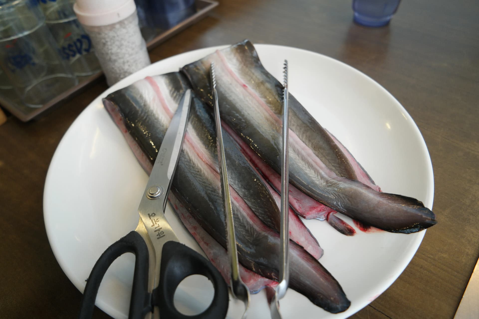 흰 접시 위에 펼쳐진 손질된 민물장어 날것 모습 - 가위와 집게가 함께 놓여 있다 | 하이제이에스비