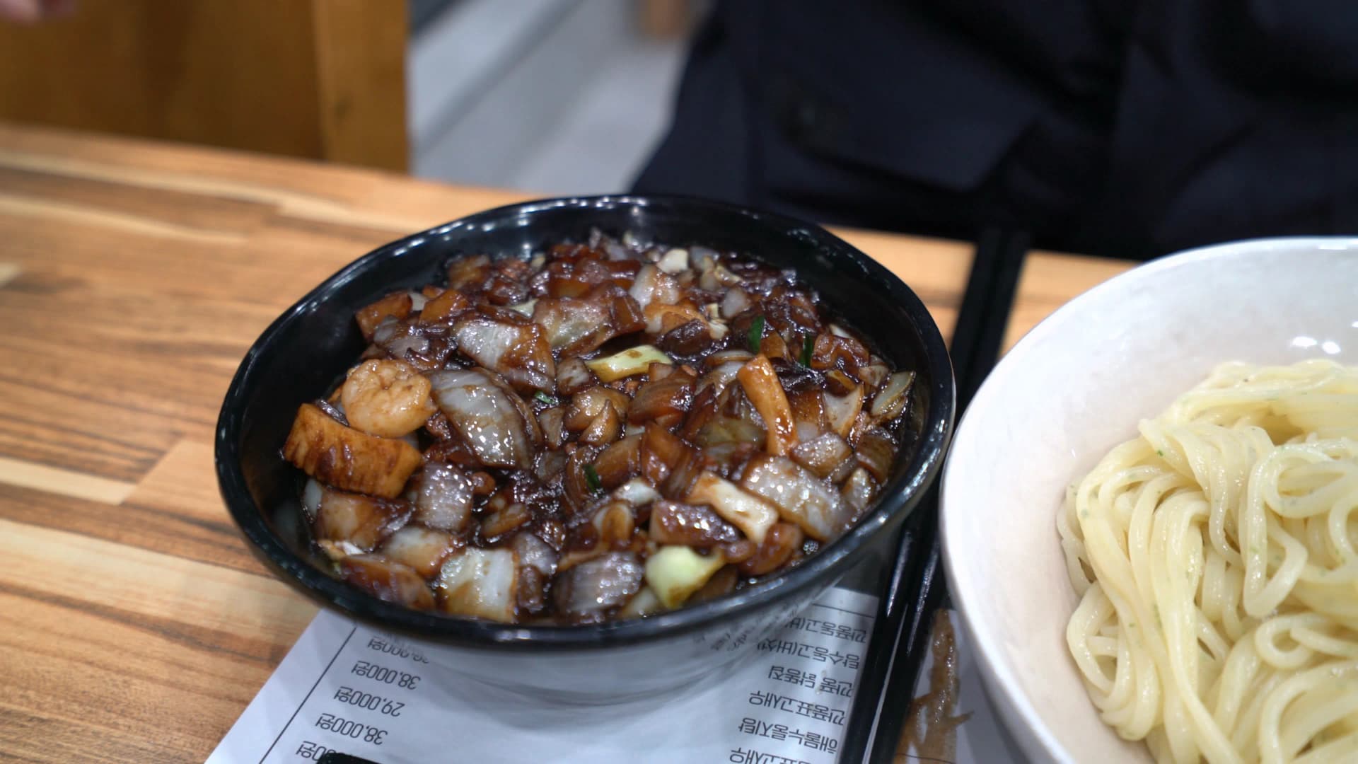 وعاء صوص كانجاجانغ أسود لامع بجانب وعاء نودلز أبيض على الطاولة، التباين بين السواد والبياض واضح