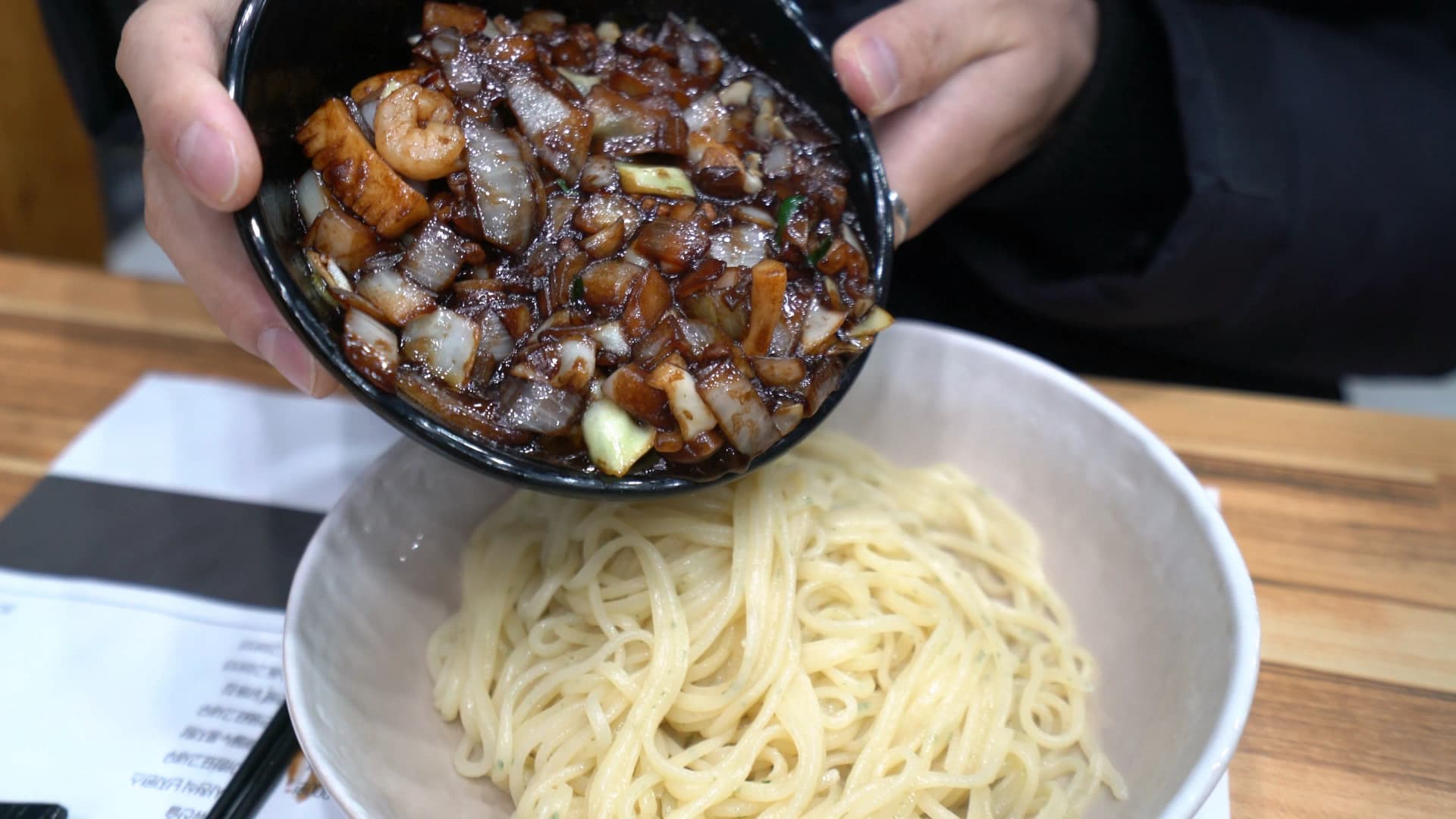 لحظة سكب صوص الكانجاجانغ من الوعاء الأسود فوق النودلز البيضاء، الصوص الداكن ينسكب على الشعيرات