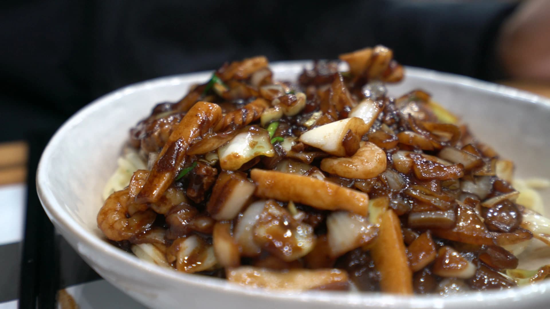 نودلز كانجاجانغ بعد سكب كل الصوص، قطع بصل ولحم وحبّار مكدّسة فوق النودلز والصوص الأسود يلمع بالزيت