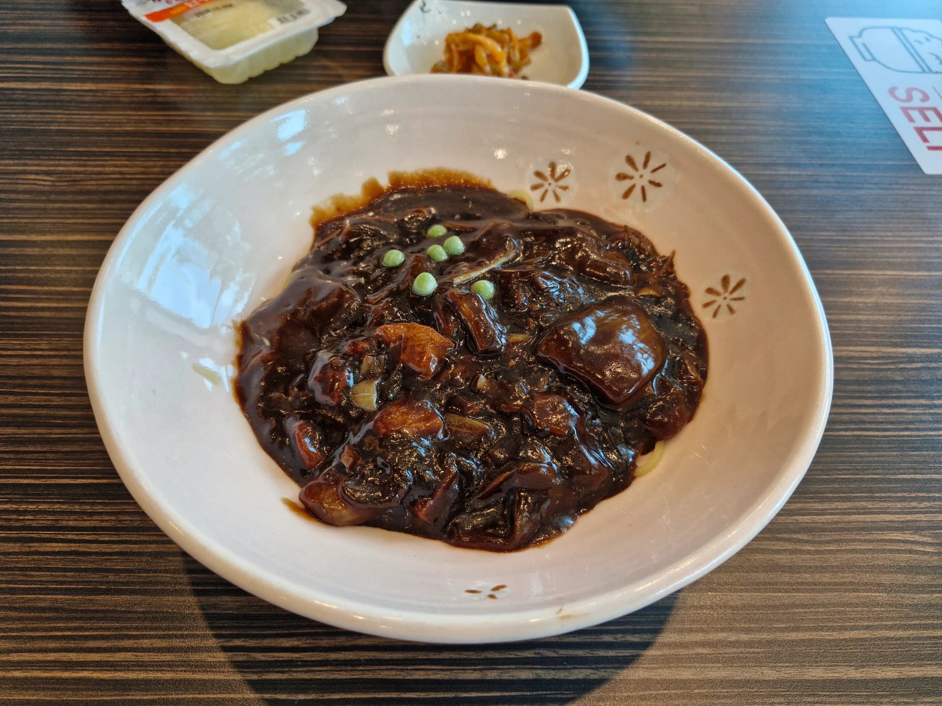 طبق جاجانغميون كلاسيكي في وعاء مزخرف بأزهار الكرز، صوص أسود داكن من معجون التشونجانغ يغطّي النودلز مع حبّات بازلاء خضراء، وبجانبه دانموجي مخلّل وبصل مخلّل