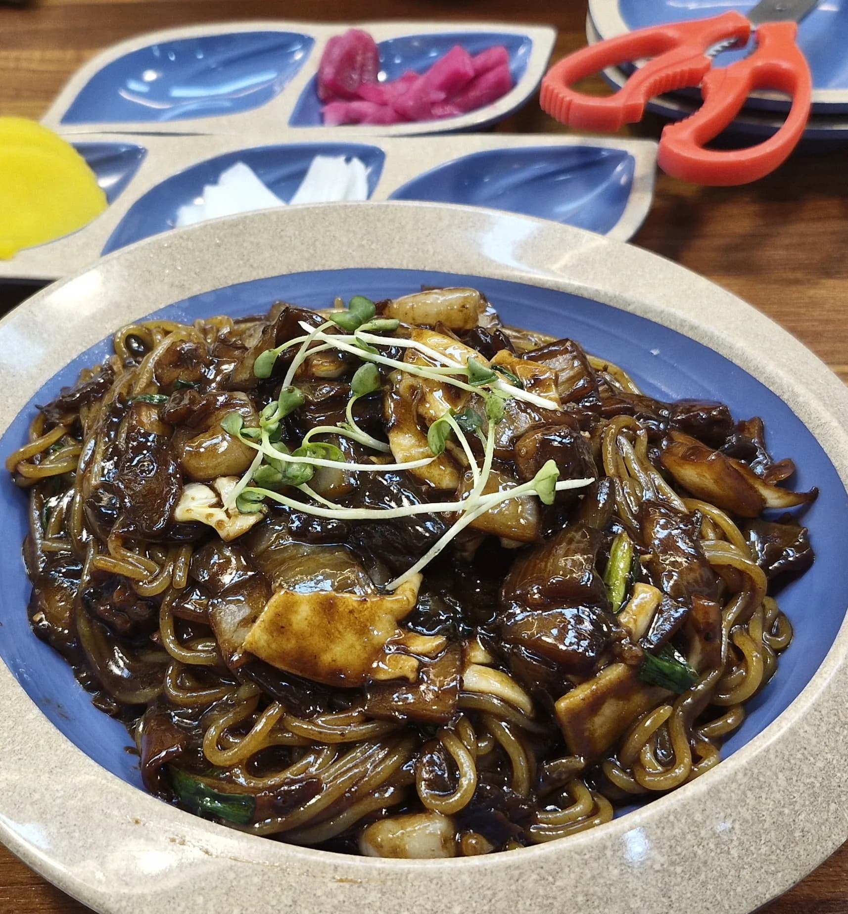 لقطة مقرّبة لقطع حبّار وروبيان مغلّفة بصوص التشونجانغ الأسود، وفي الخلفية صحن مقسّم فيه دانموجي وبصل مخلّل وزنجبيل وردي