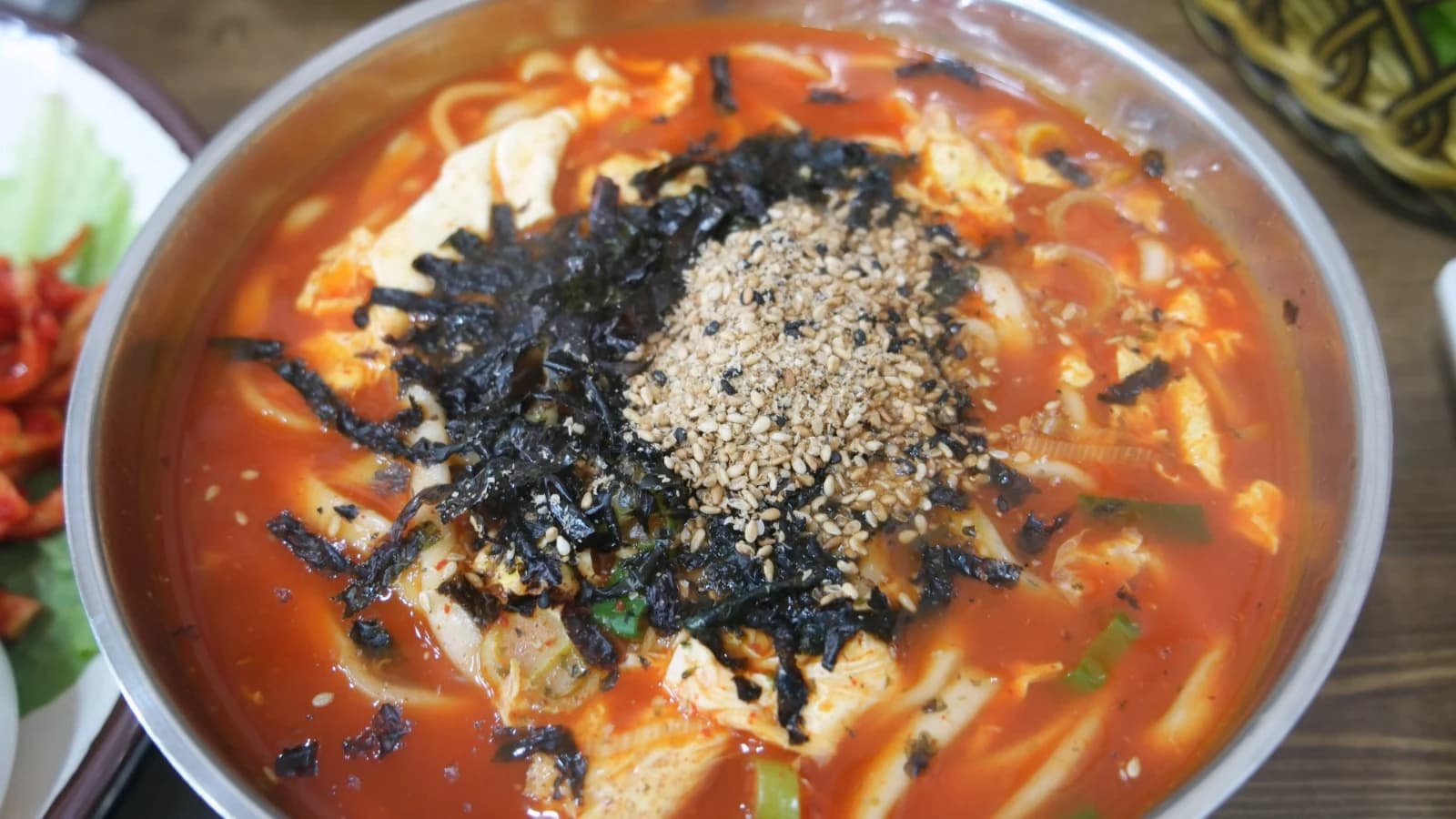 Close-up of spicy kalguksu broth with scattered seaweed flakes and a mound of sesame seeds