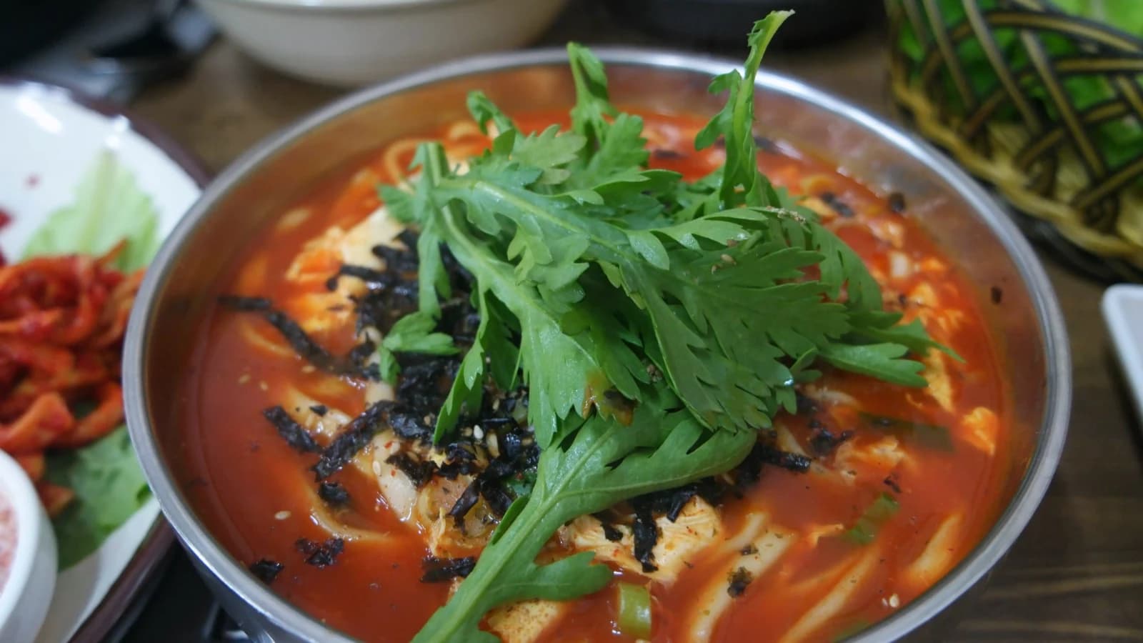 Handful of crown daisy greens piled on top of spicy kalguksu contrasting green leaves against red broth