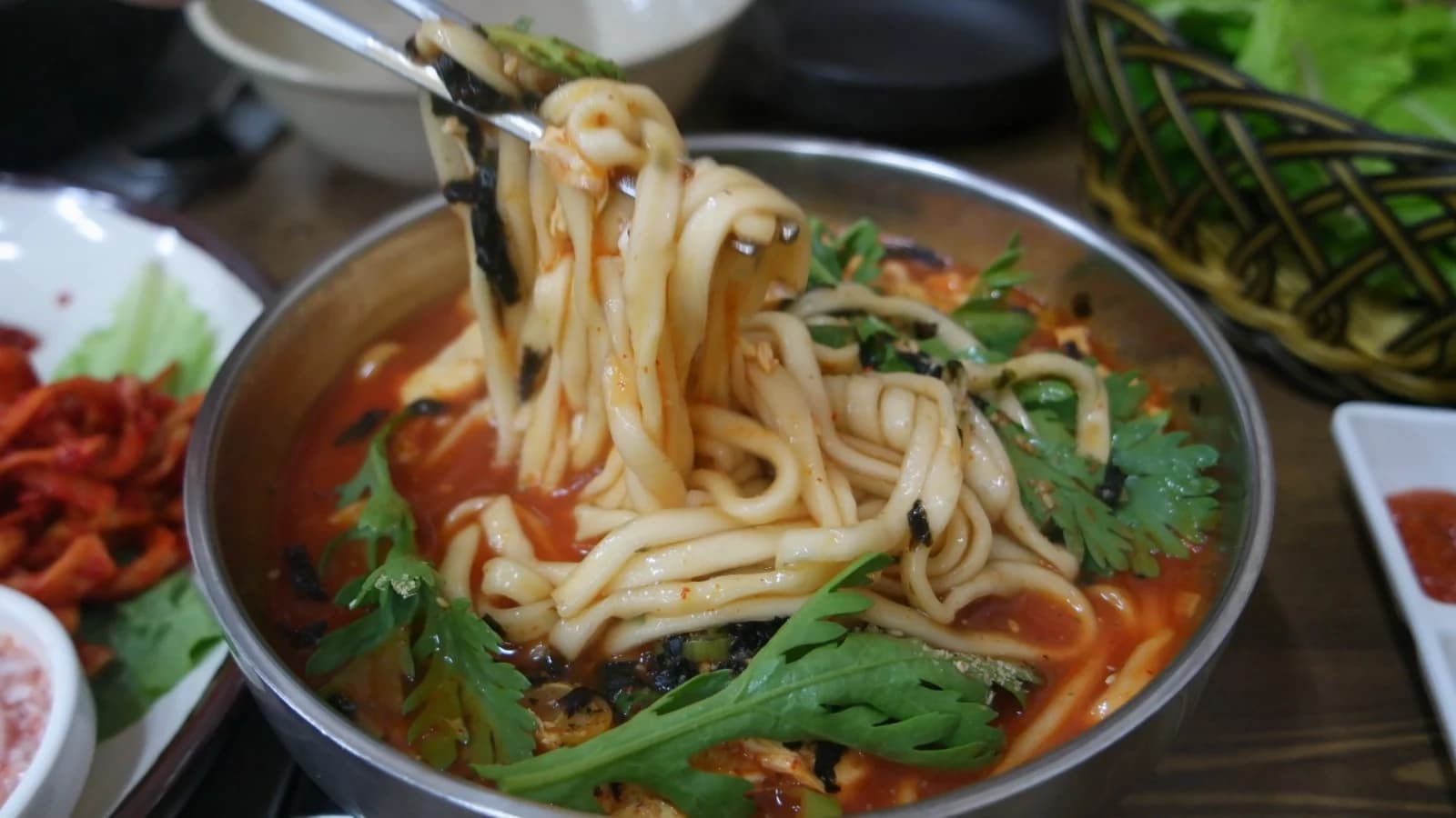 Chopsticks lifting hand-cut kalguksu noodles of varying thickness from spicy red broth