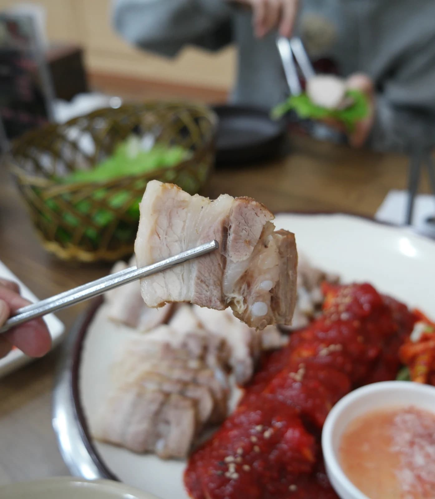 Chopsticks lifting a slice of bossam pork with someone wrapping a lettuce wrap in the background