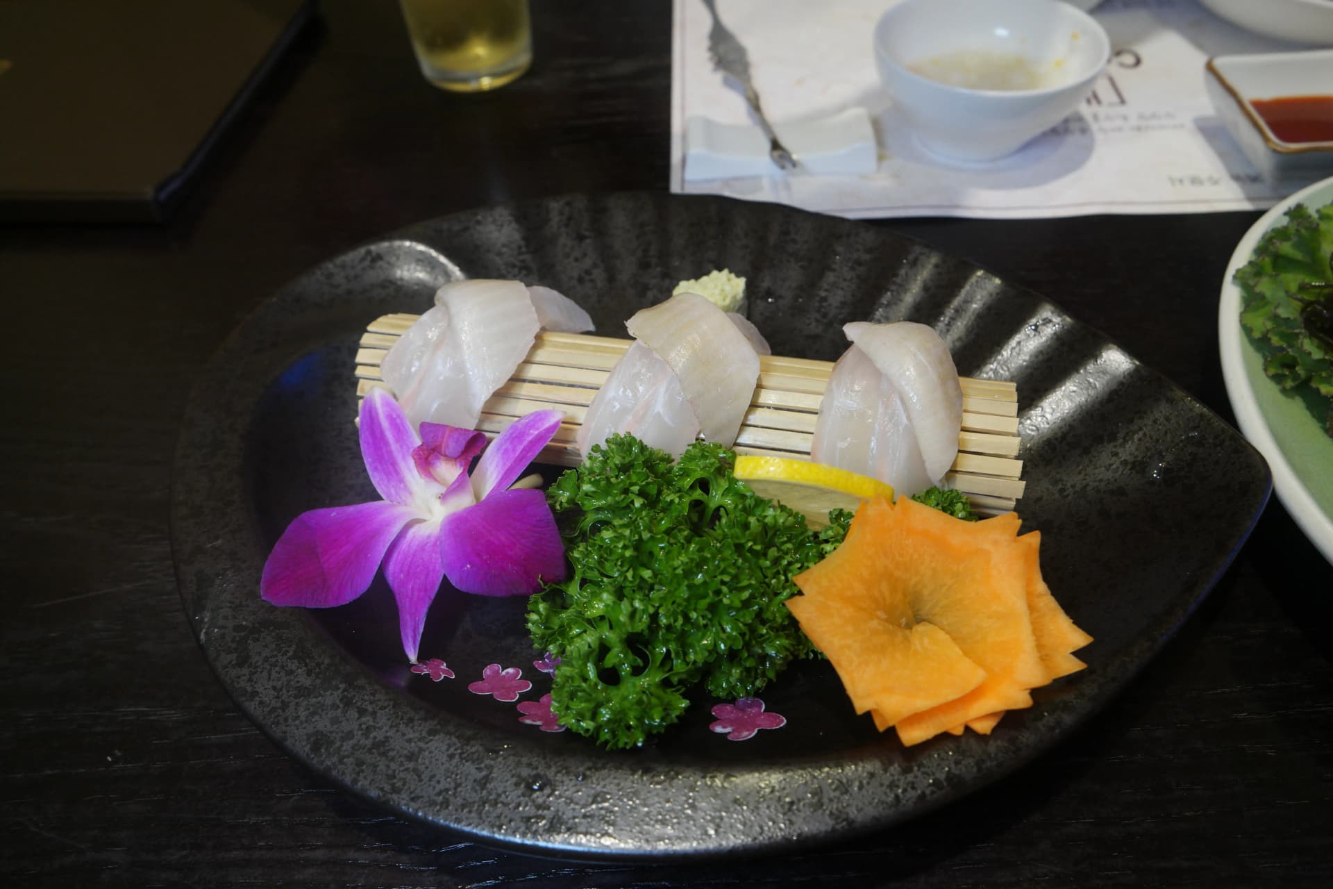 Lonchas gruesas de sashimi de lenguado coreano presentadas sobre una esterilla de bambú con orquídea, zanahoria y limón como guarnición | 하이제이에스비