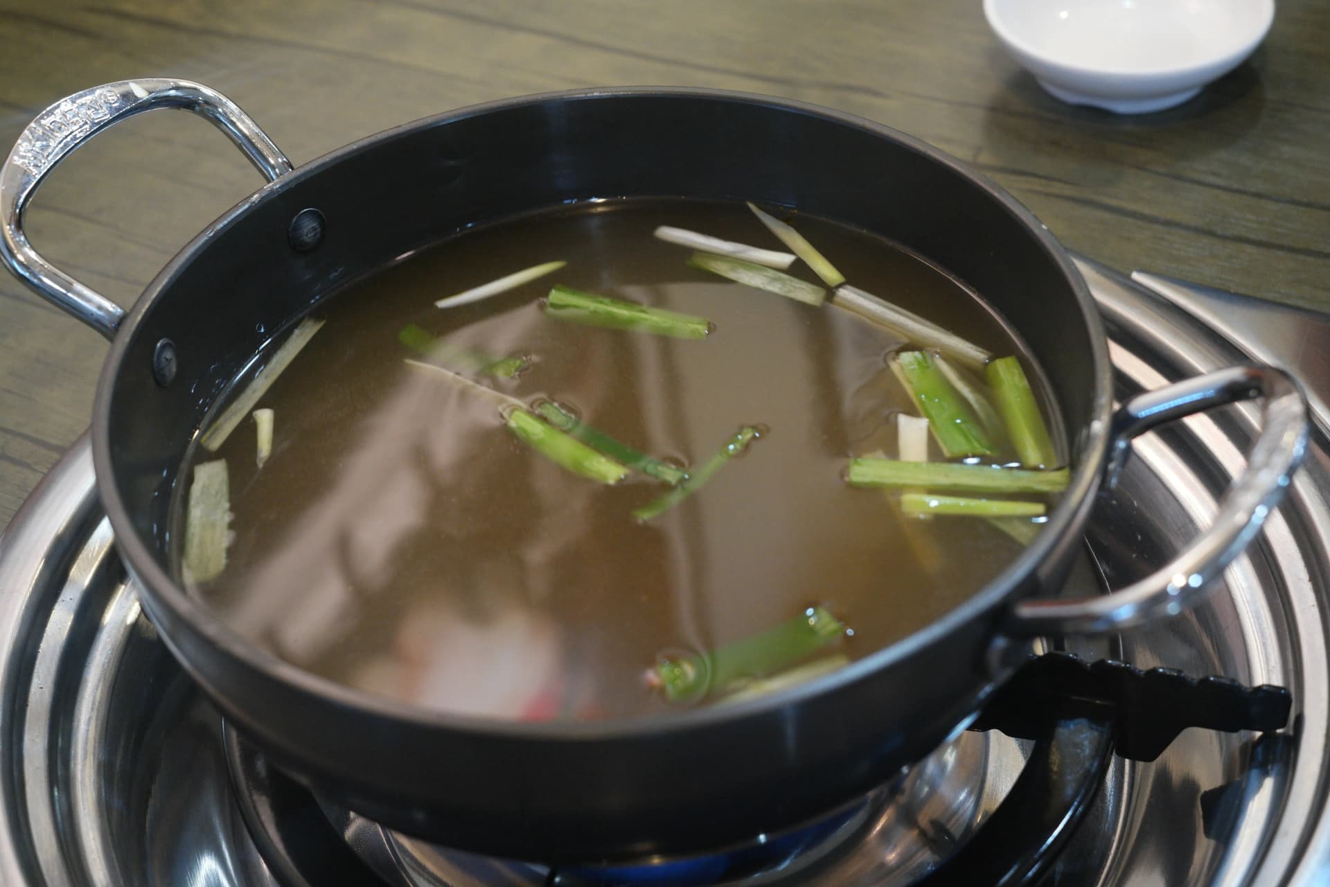 테이블 위 버너에 올려진 전골 냄비에 맑은 육수와 대파가 담겨 있는 모습 | 하이제이에스비