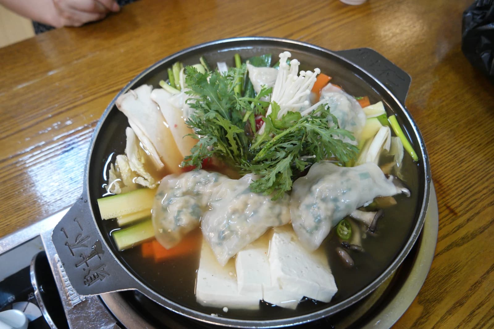 Mandu jeongol overview — handmade dumplings, tofu, enoki mushrooms, crown daisy greens, and carrots arranged in a wide hot pot filled with soy-based broth