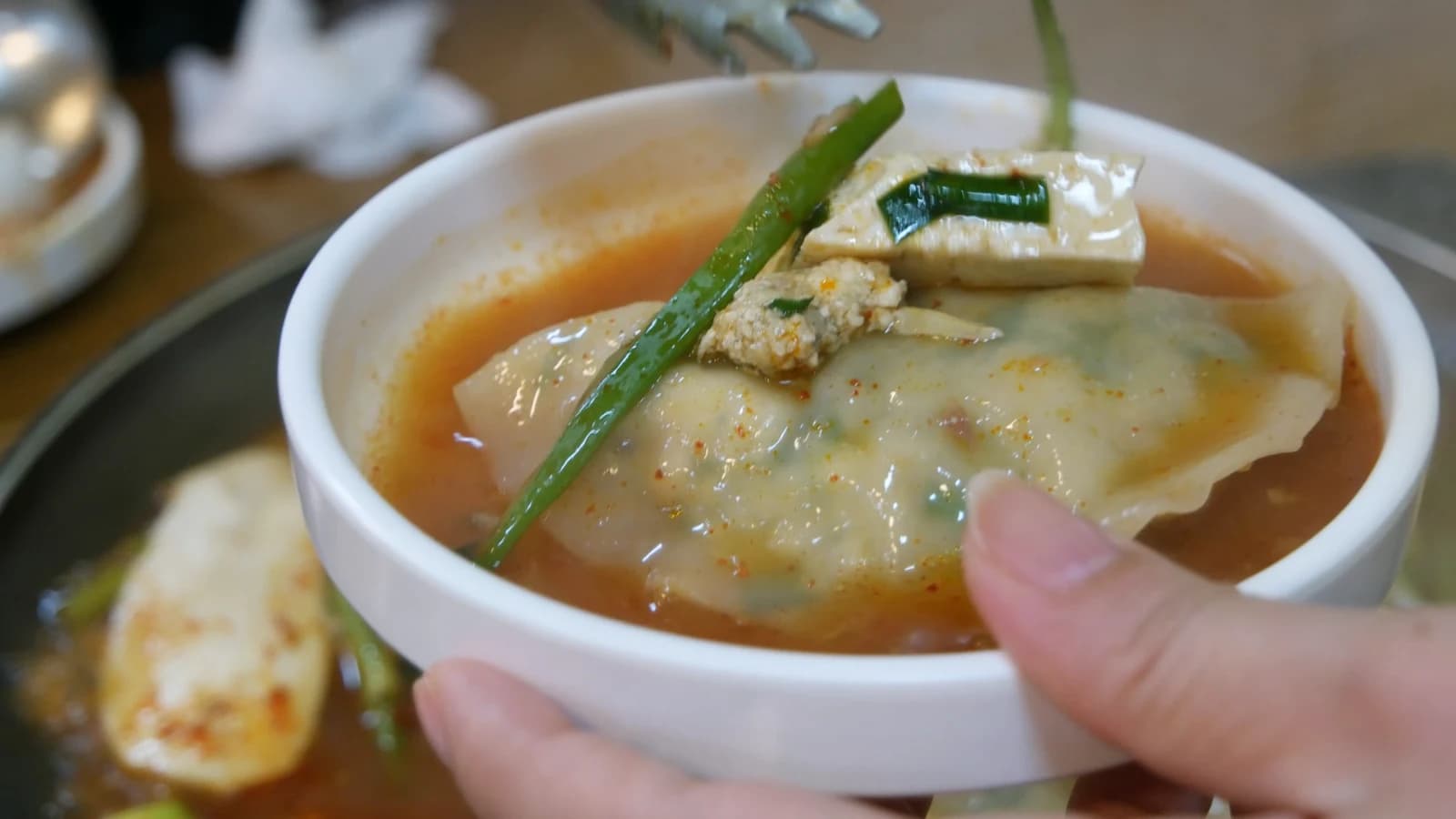 How to eat mandu jeongol — small bowl held in hand with dumpling, tofu, and rich dark broth ladled out from the hot pot