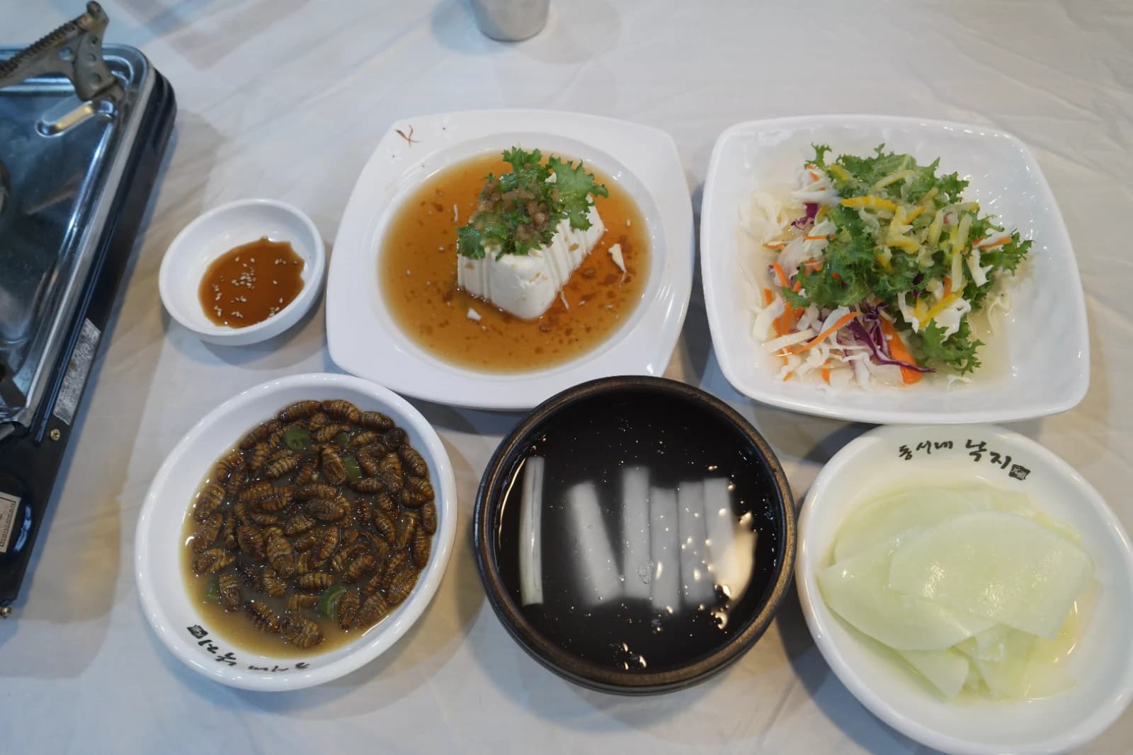 Full table setup of banchan side dishes at a Korean octopus and intestine stir-fry restaurant including silken tofu salad and dumplings