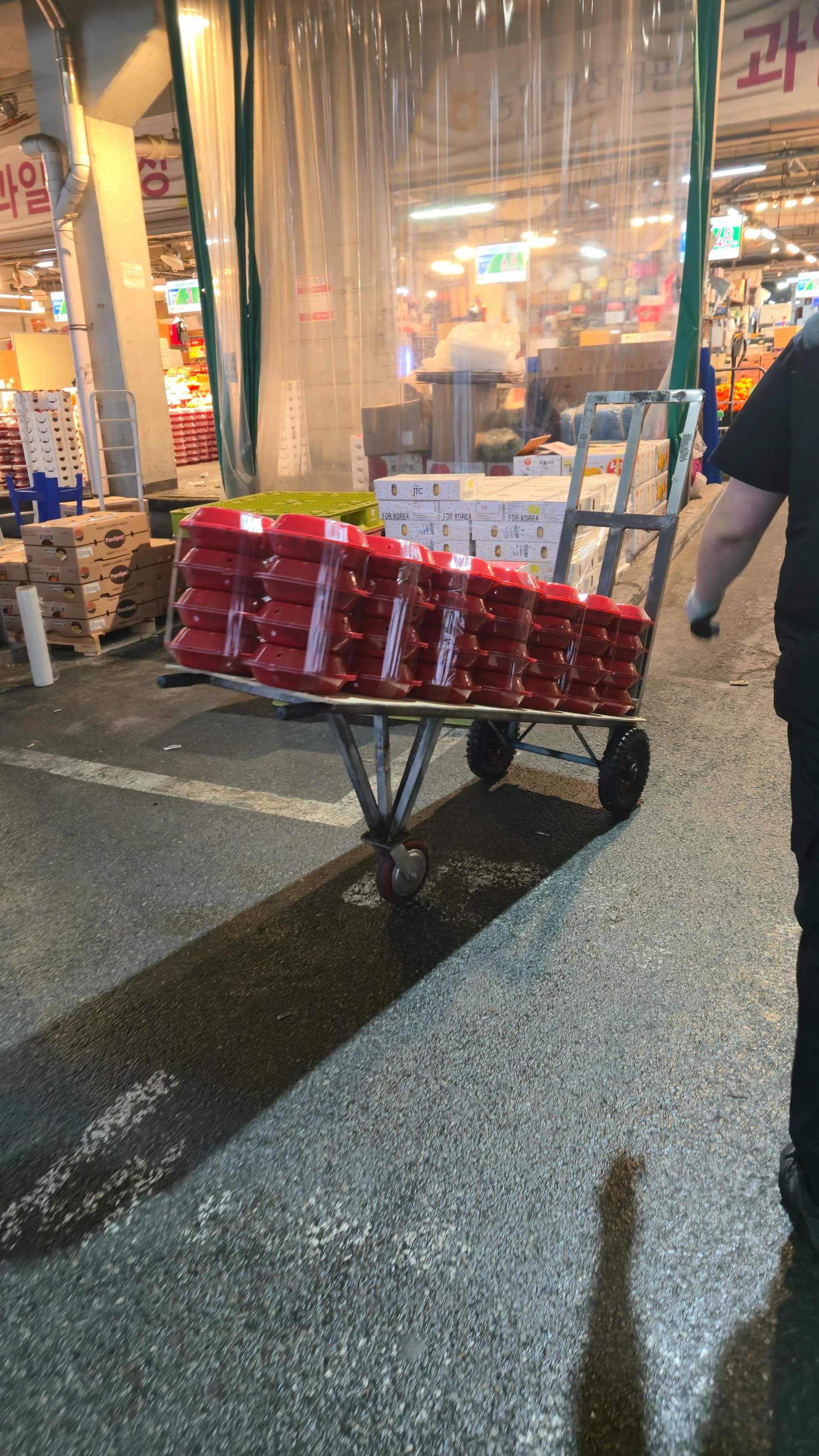 시장 입구에서 빨간 포장 용기가 실린 수레가 지나가는 모습, 상인들이 물건을 옮기고 있다