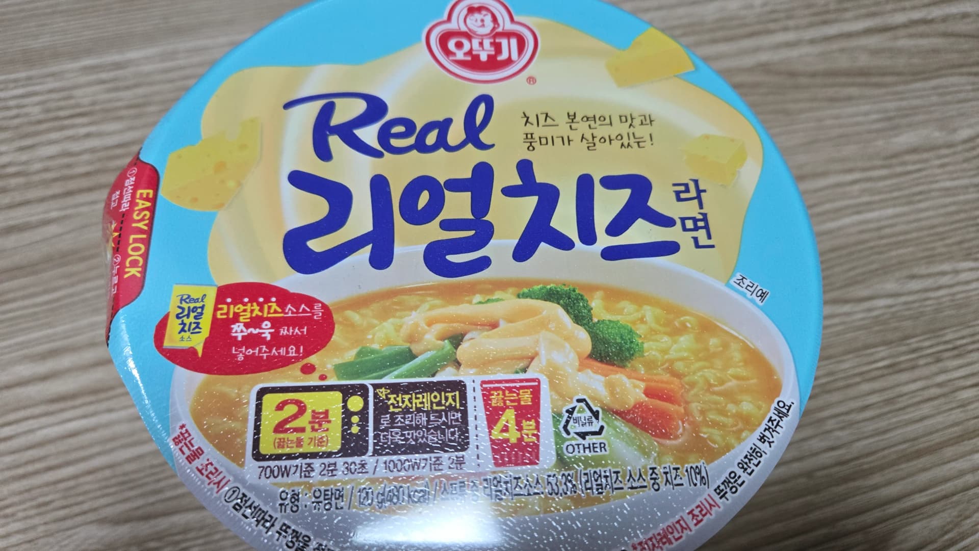 Part frontal del got de ramen de formatge d’Ottogi — envàs gran amb combinació de blau cel i groc | 하이제이에스비