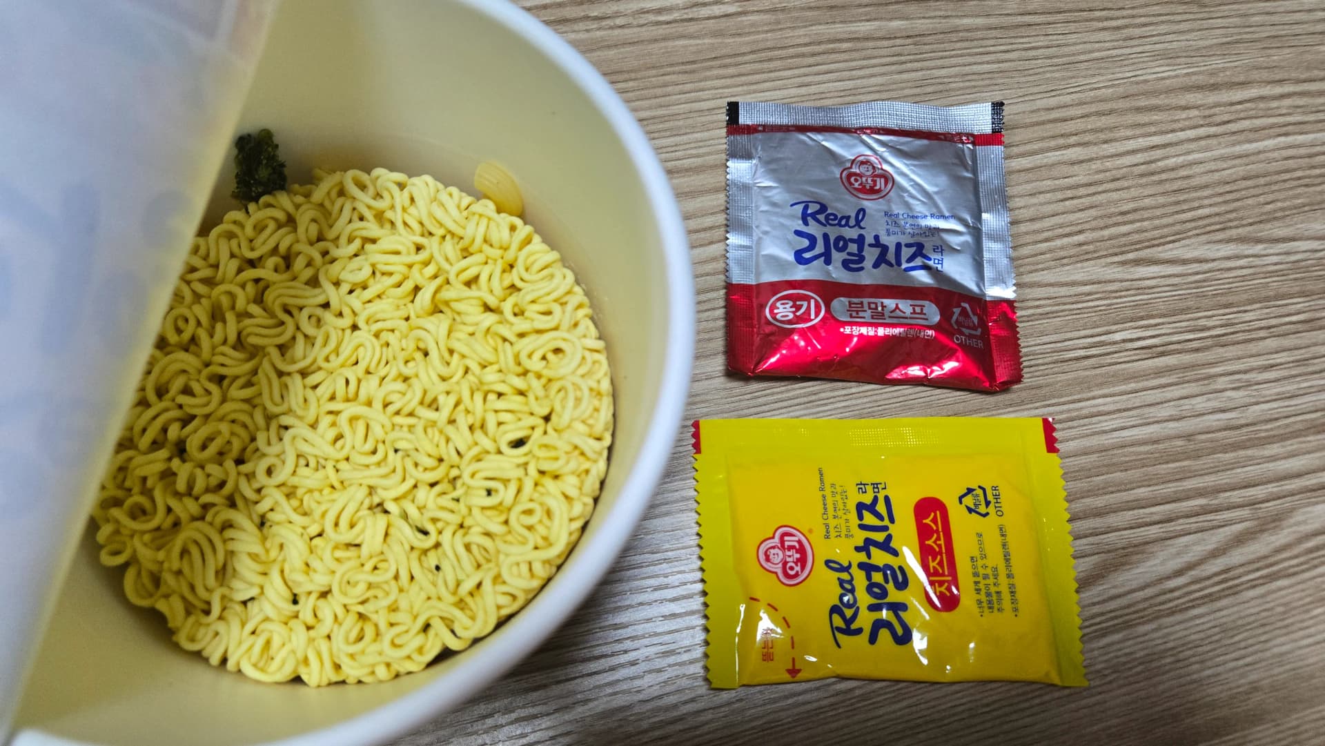 Contingut del got de ramen de formatge d’Ottogi — fideus grocs, sobre vermell de sopa en pols i sobre groc de salsa Real Cheese | 하이제이에스비