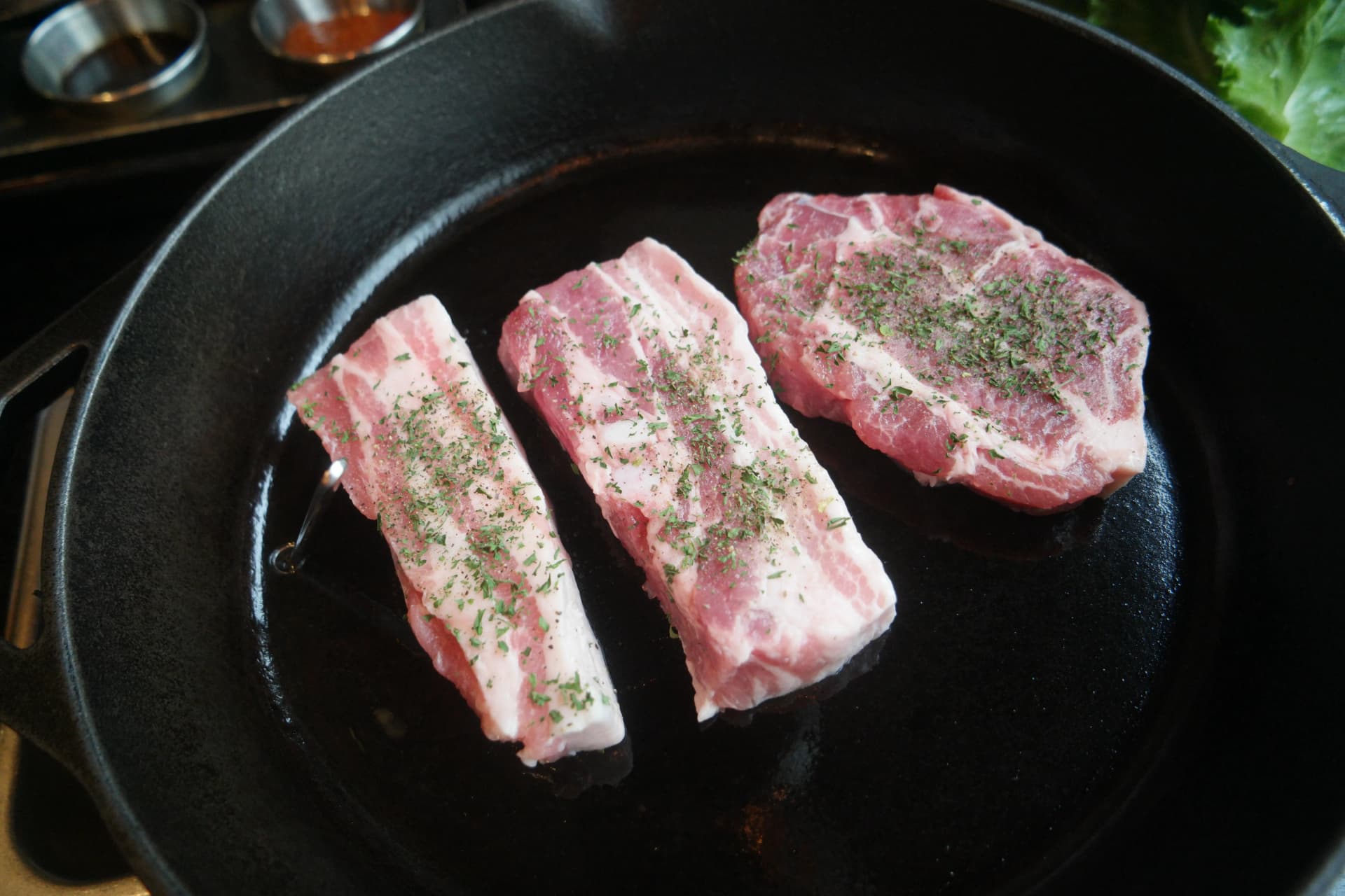 Due fette di pancetta e una di spalla di maiale disposte sulla padella in ghisa calda | 하이제이에스비
