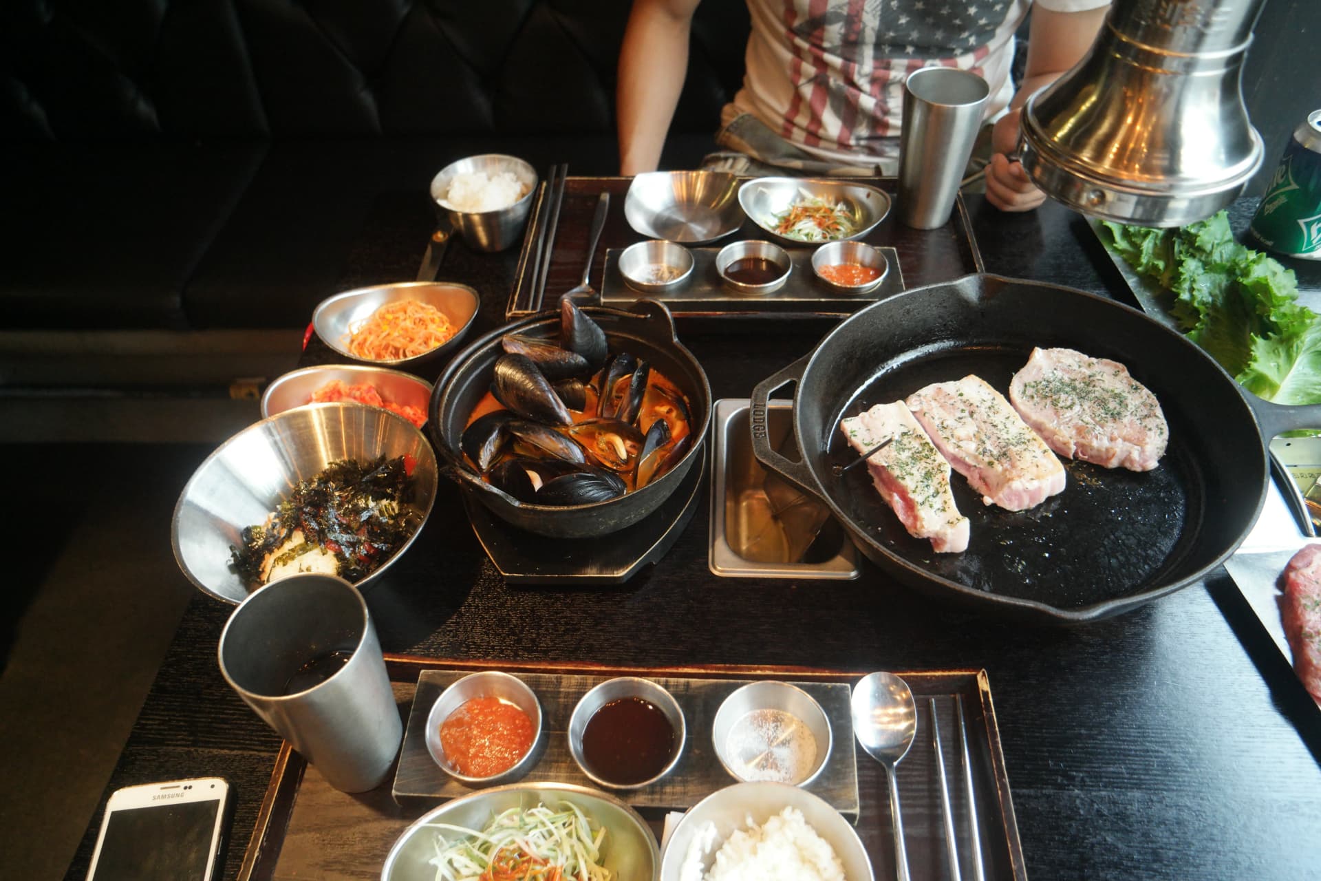Visione d'insieme del tavolo in un ristorante coreano a volontà con padella in ghisa, zuppa di cozze e vari contorni | 하이제이에스비