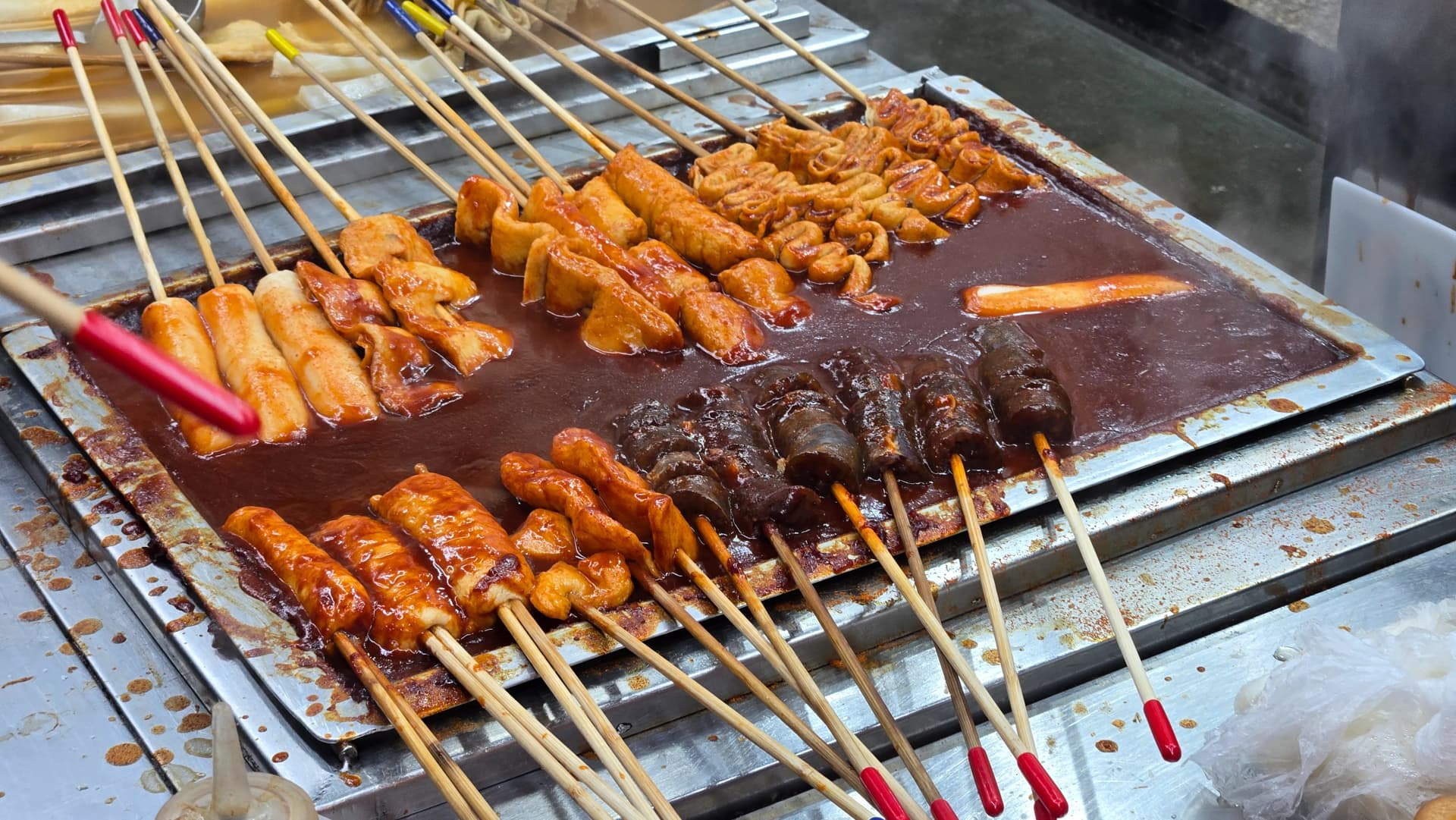 Gochujang-Spieße auf dem Seomun-Markt in Daegu — breite Grillplatte voll mit Reiskuchen-, Fischkuchen- und Sundae-Spießen in roter Chilipaste | 하이제이에스비