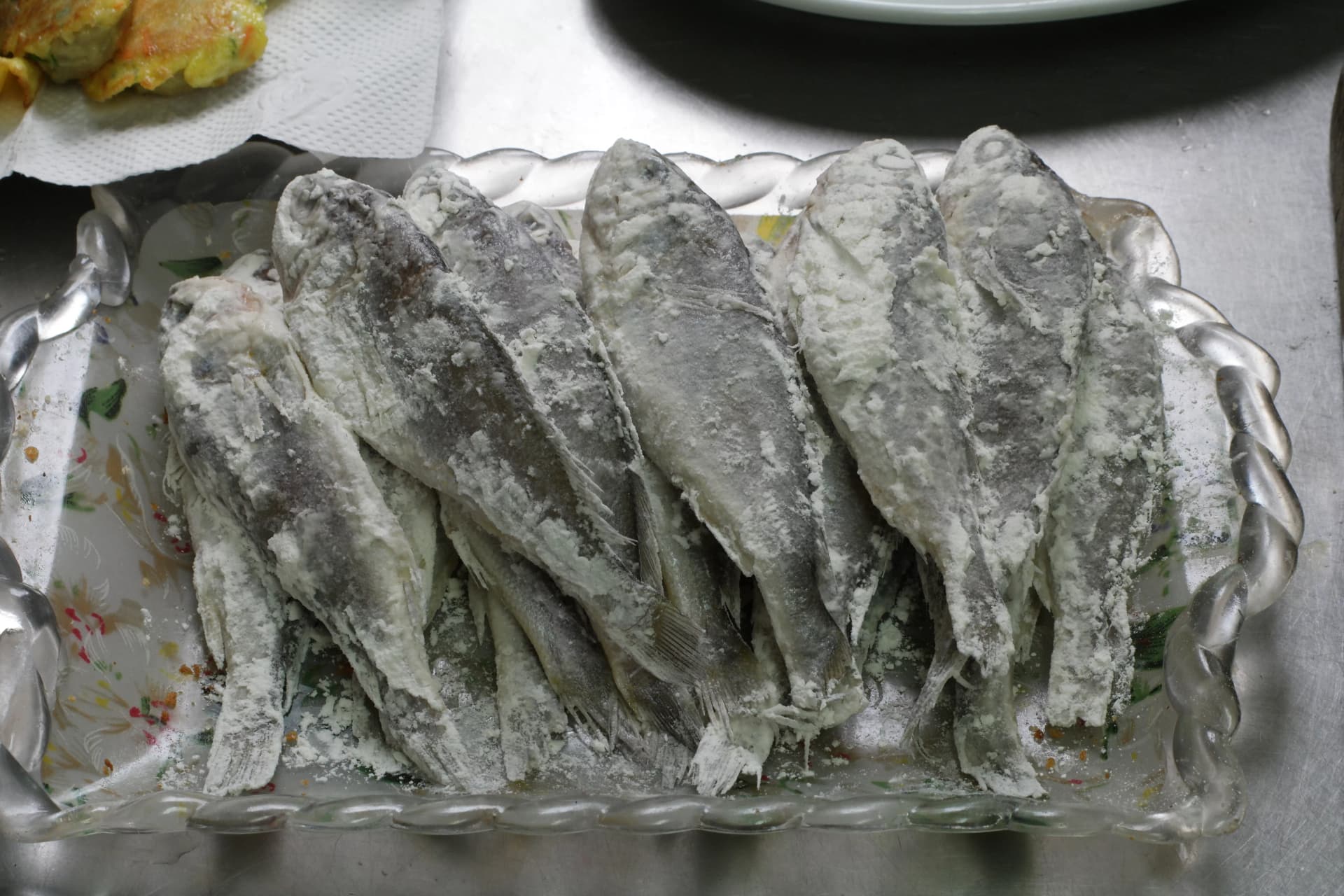 Several flour-coated yellow croaker fish lined up on a glass plate before frying, a classic Korean home-style side dish