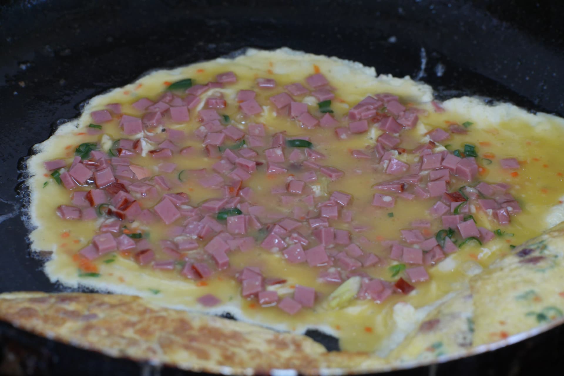Thin layer of egg mixture spread across a dark frying pan with visible ham and vegetables cooking for gyeran-mari