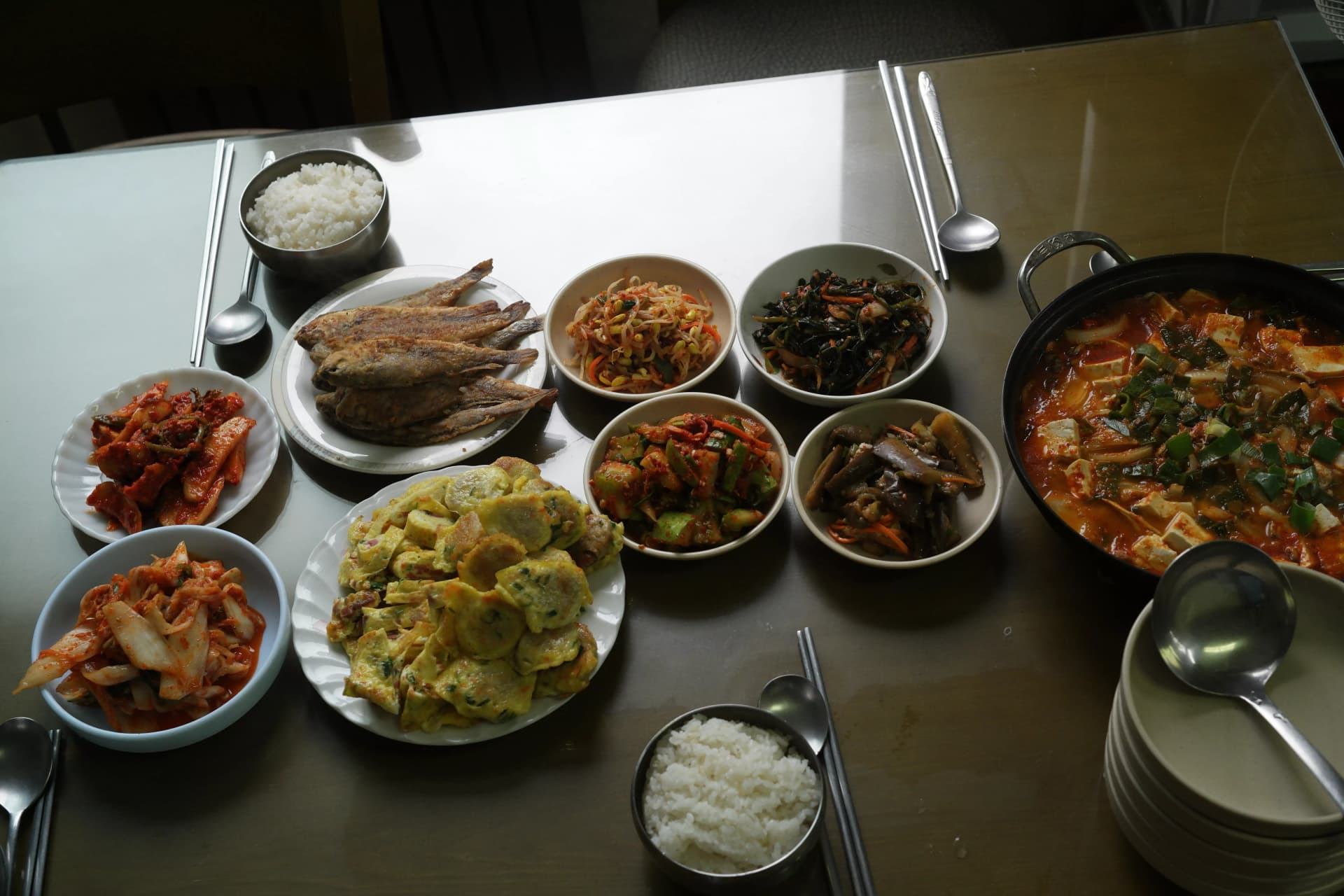 Complete Korean baekban table setting on stainless steel surface with rice kimchi jjigae pan-fried croaker rolled omelette meat patties and assorted banchan side dishes