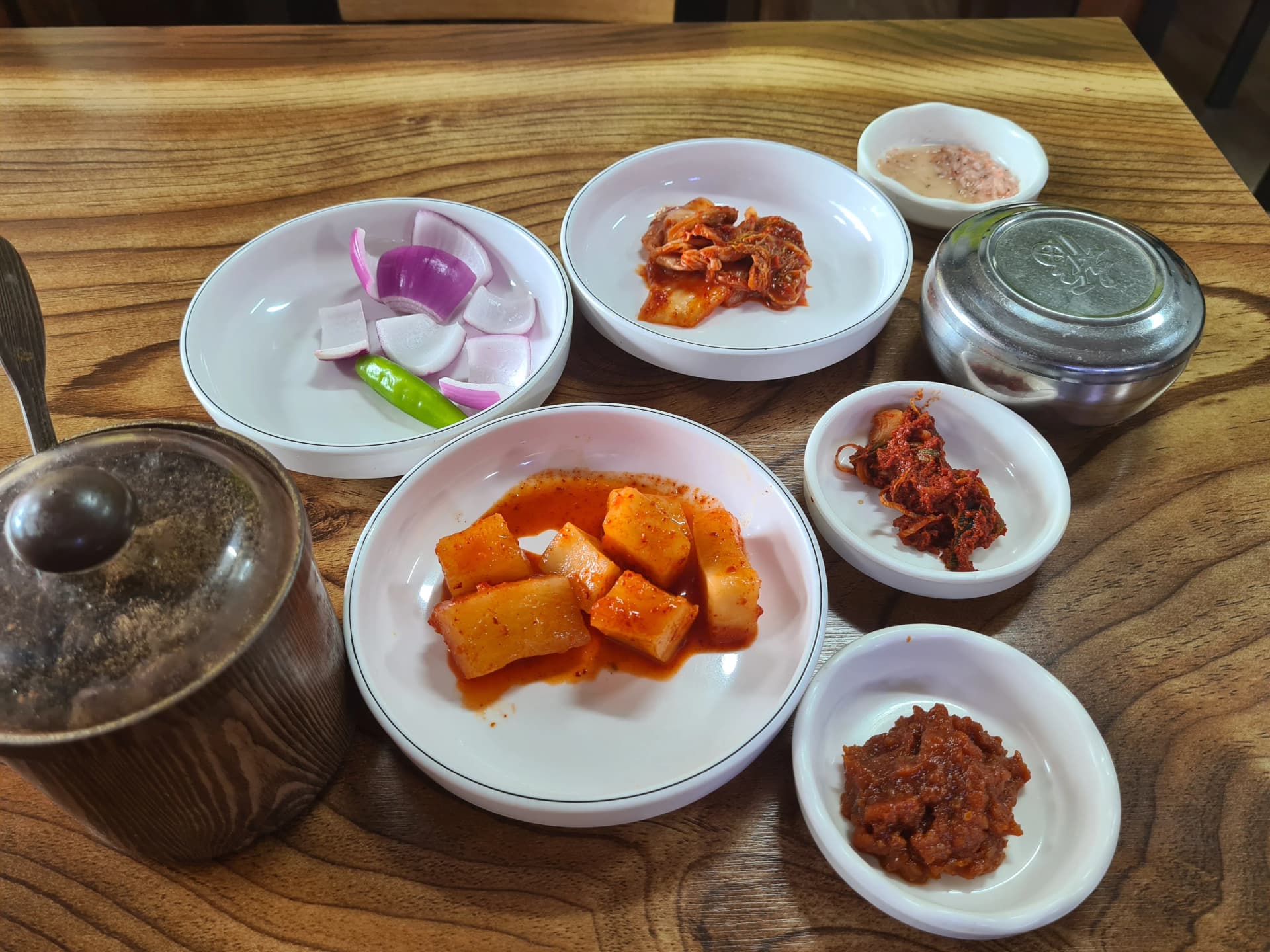 ترتيب المقبلات الجانبية بمطعم سوندي غوكباب — كاكدوقي وكيمتشي ملفوف وبصل وفلفل أخضر حار ومعجون فول صويا وروبيان مملح وإبريق مرق على طاولة خشبية