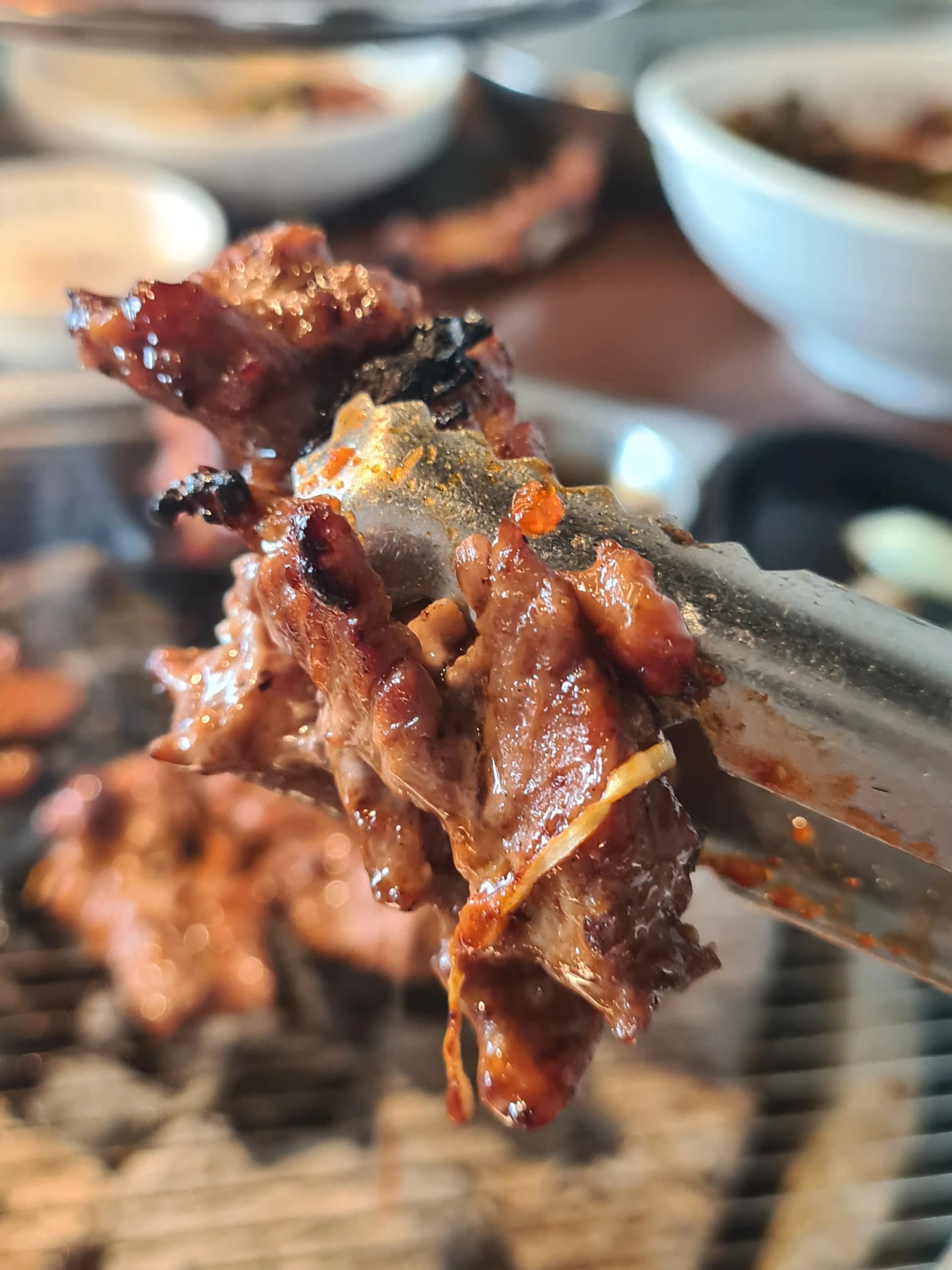 ريش لحم متبّلة مكتملة الشوي مرفوعة بالملقط — سطح مكرمل ومدخّن بنكهة الفحم